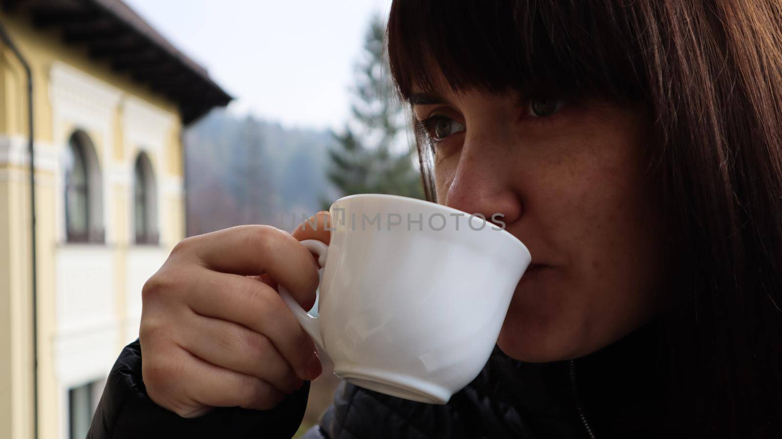 Beautiful woman with a cup of coffee and tea enjoying beautiful views of the mountain landscape from the hotel balcony. A young happy girl is having breakfast on the balcony of her hotel room. by Roshchyn