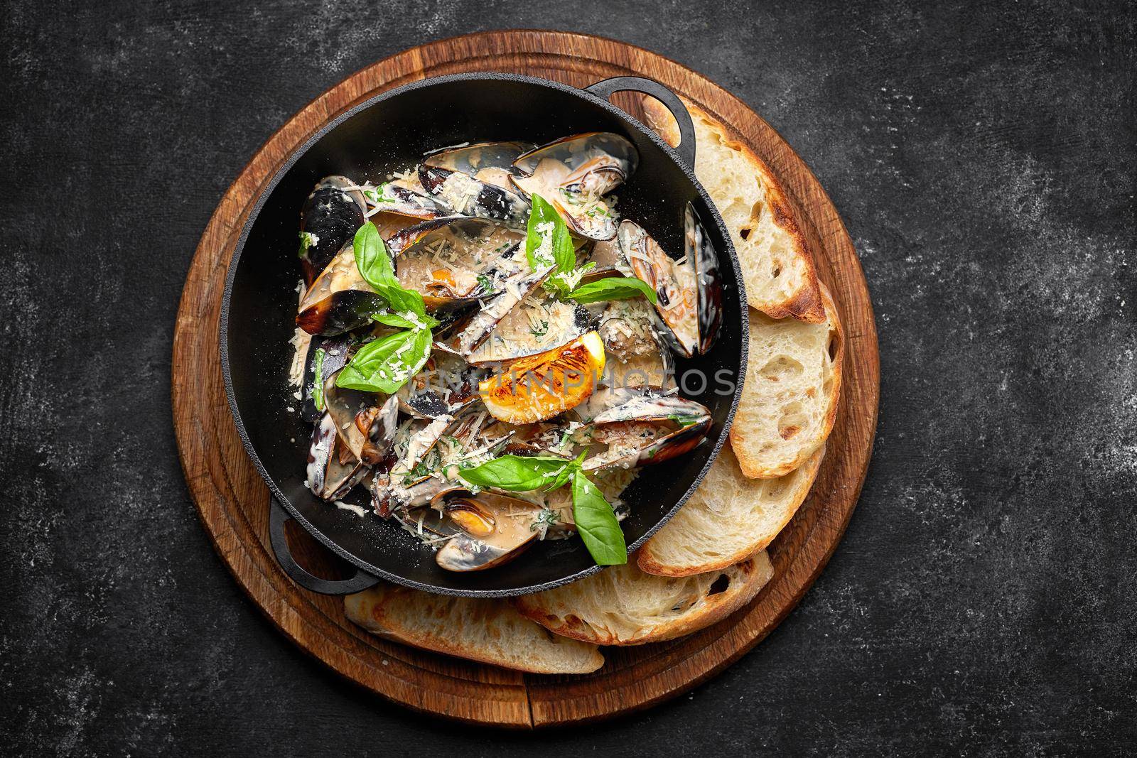 Cooked mussels in a skillet with cheese and basil leaves, on a wooden board, on a dark background