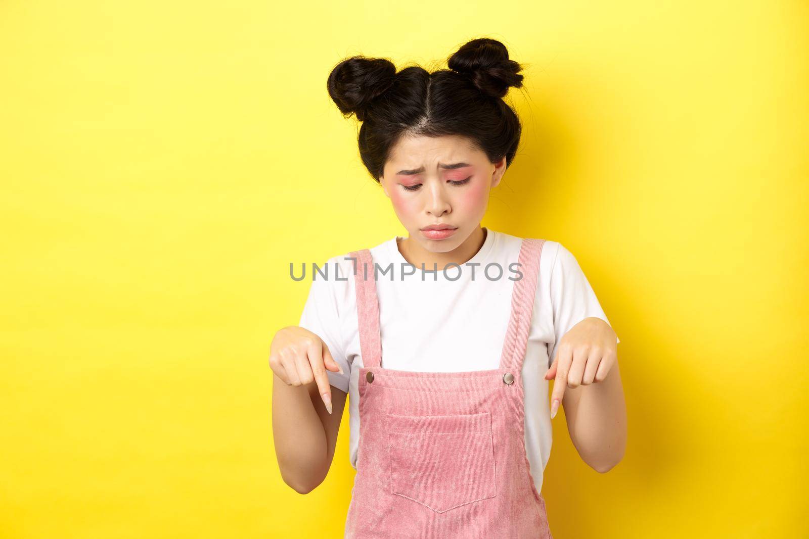 Sad miserable girl with glamour bright makeup, looking and pointing down upset, showing bad thing, standing against yellow background.