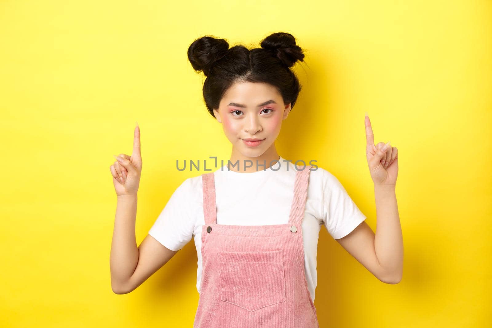 Confident asian young girl with beauty makeup, pointing fingers up, showing advertisement, standing on yellow background and smiling.