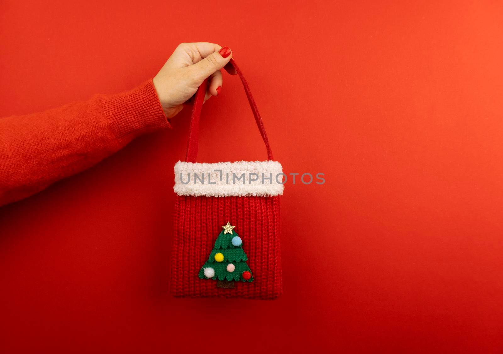Woman hand at red sweater holding red knitted Christmas bag at red background by uveita