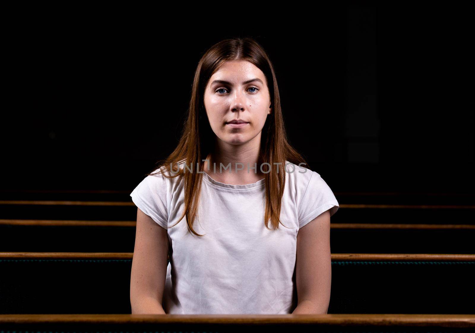 A young girl is sitting alone in a large hall looking at the camera. Around dark