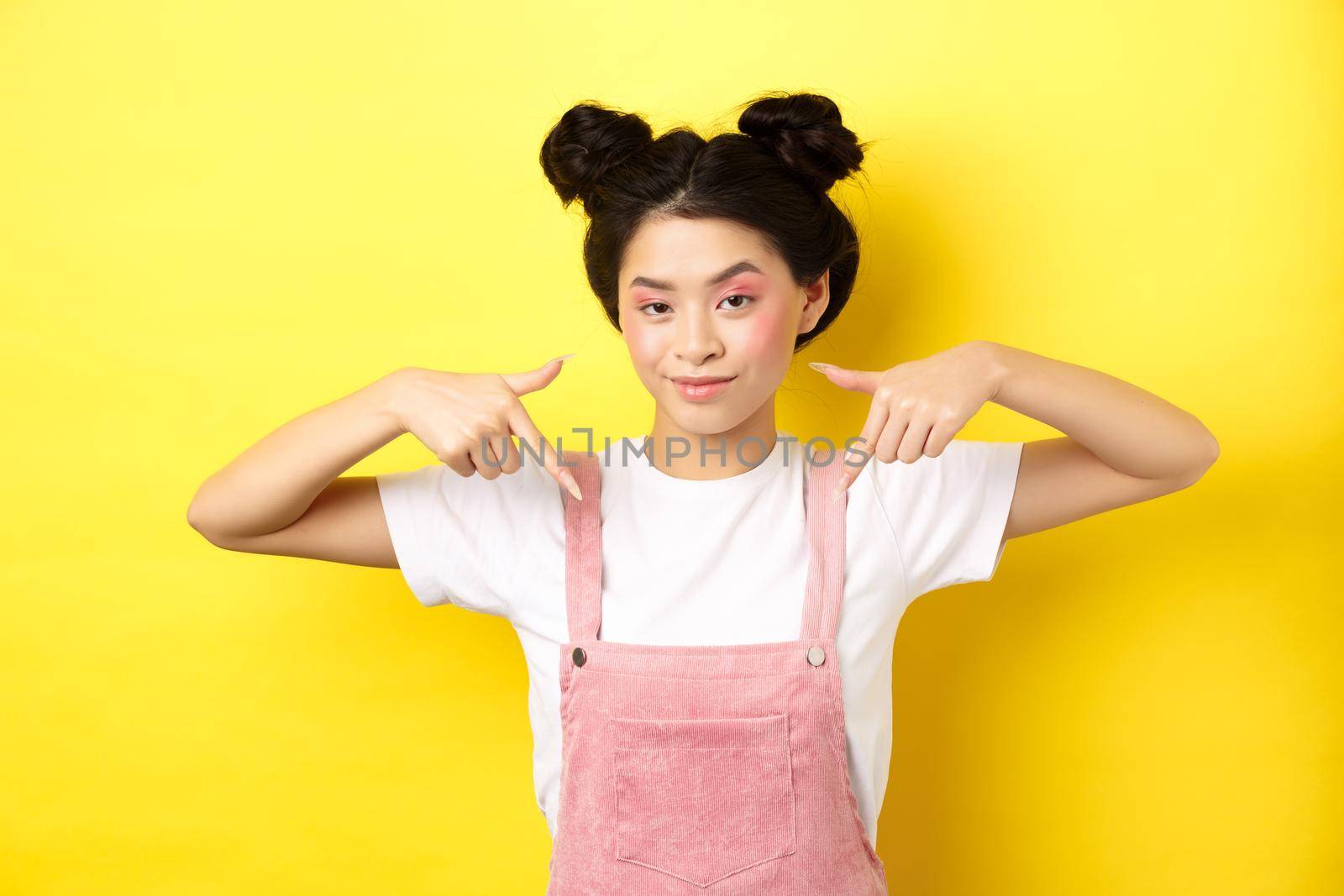 Stylish sassy girl smiling pleased, hinting on good deal, pointing fingers down at promo offer, standing in summer clothes against yellow background by Benzoix