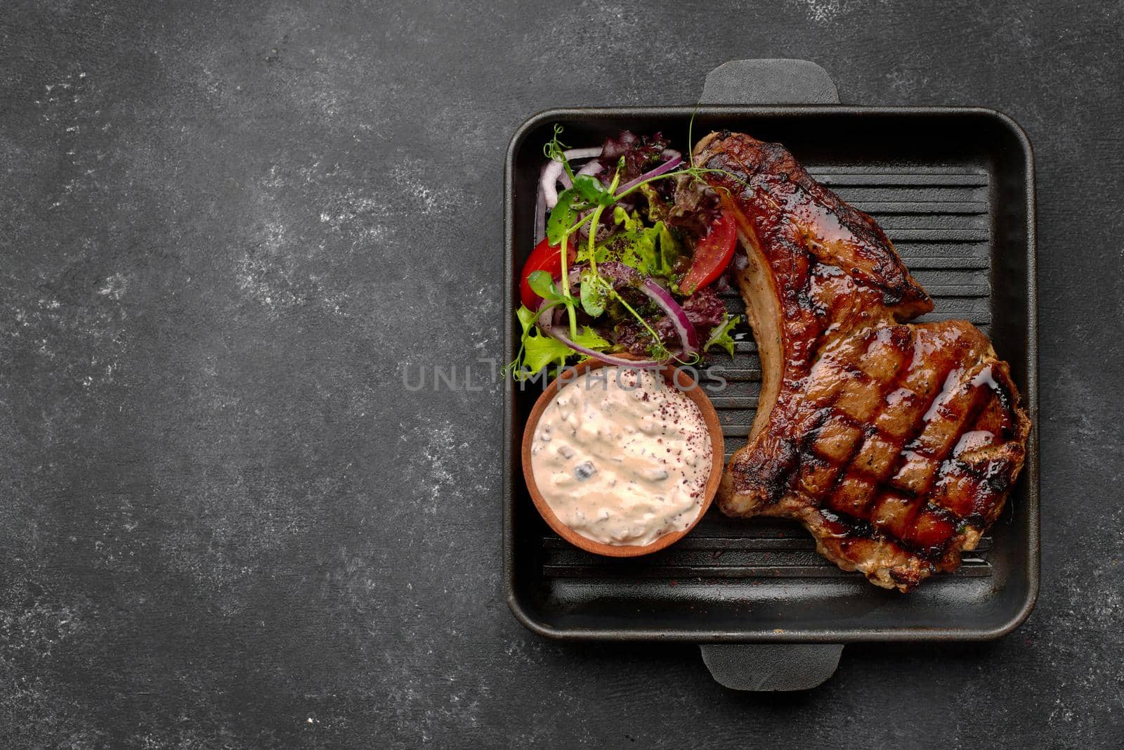 Grilled steak with sauce, vegetables and herbs, on a black frying pan, on a dark background