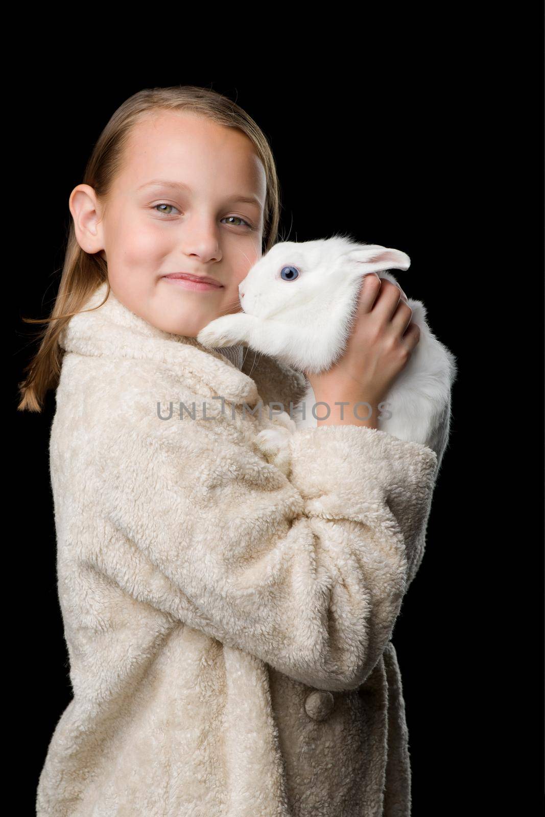 Pretty girl hugging white rabbit. Stylish beautiful girl in beige fur coat holding cute bunny. Portrait of preteen child posing with pet animal in studio against black background