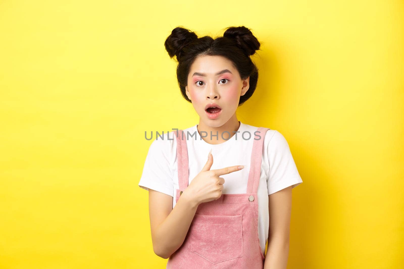 Shocked asian woman with glamour makeup, open mouth and pointing right at logo, showing advertisement, standing against yellow background by Benzoix