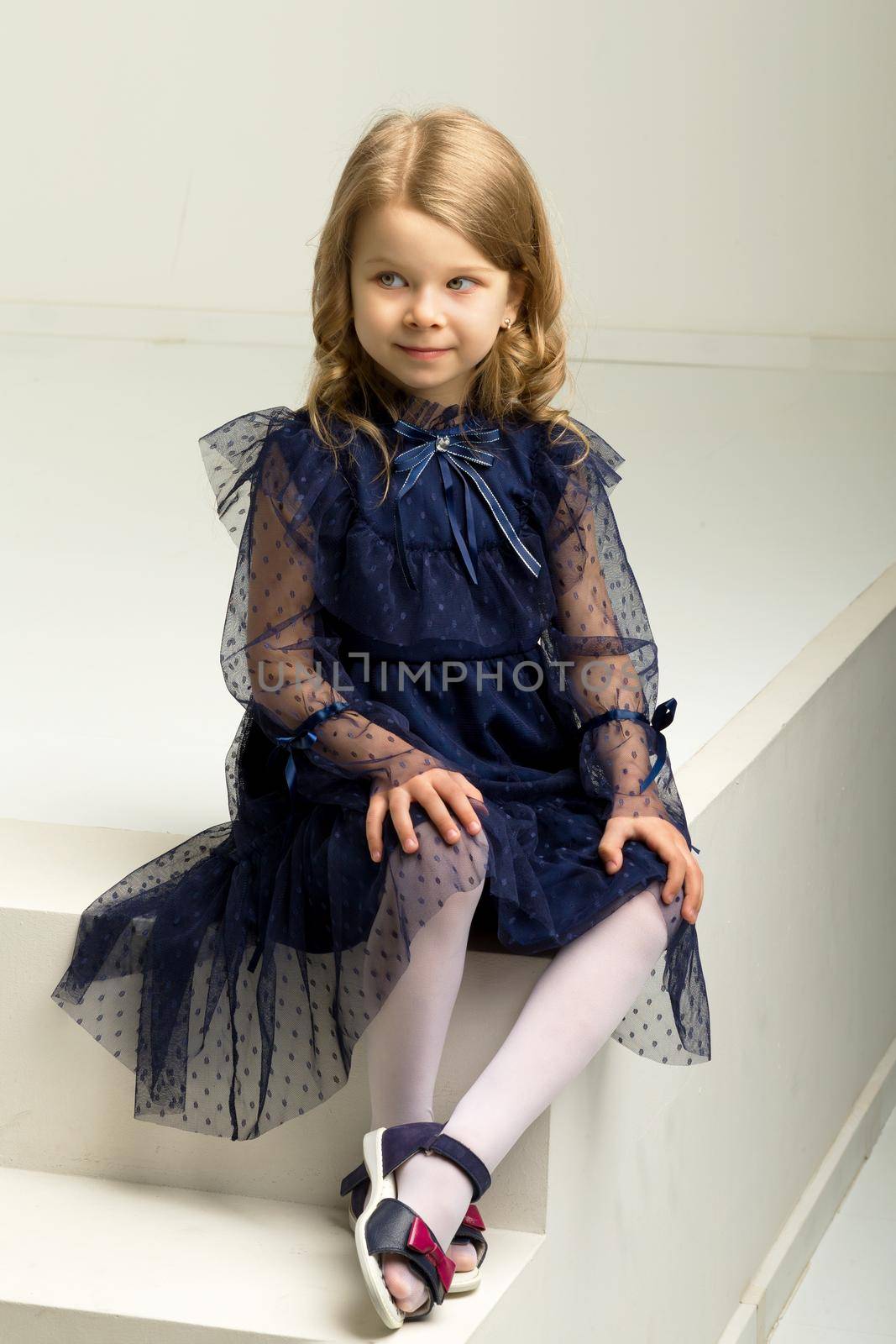 Lovely girl in a blue dress sitting on a white stairs. Portrait of a charming blonde in a beautiful lace dress sits and looks at the camera