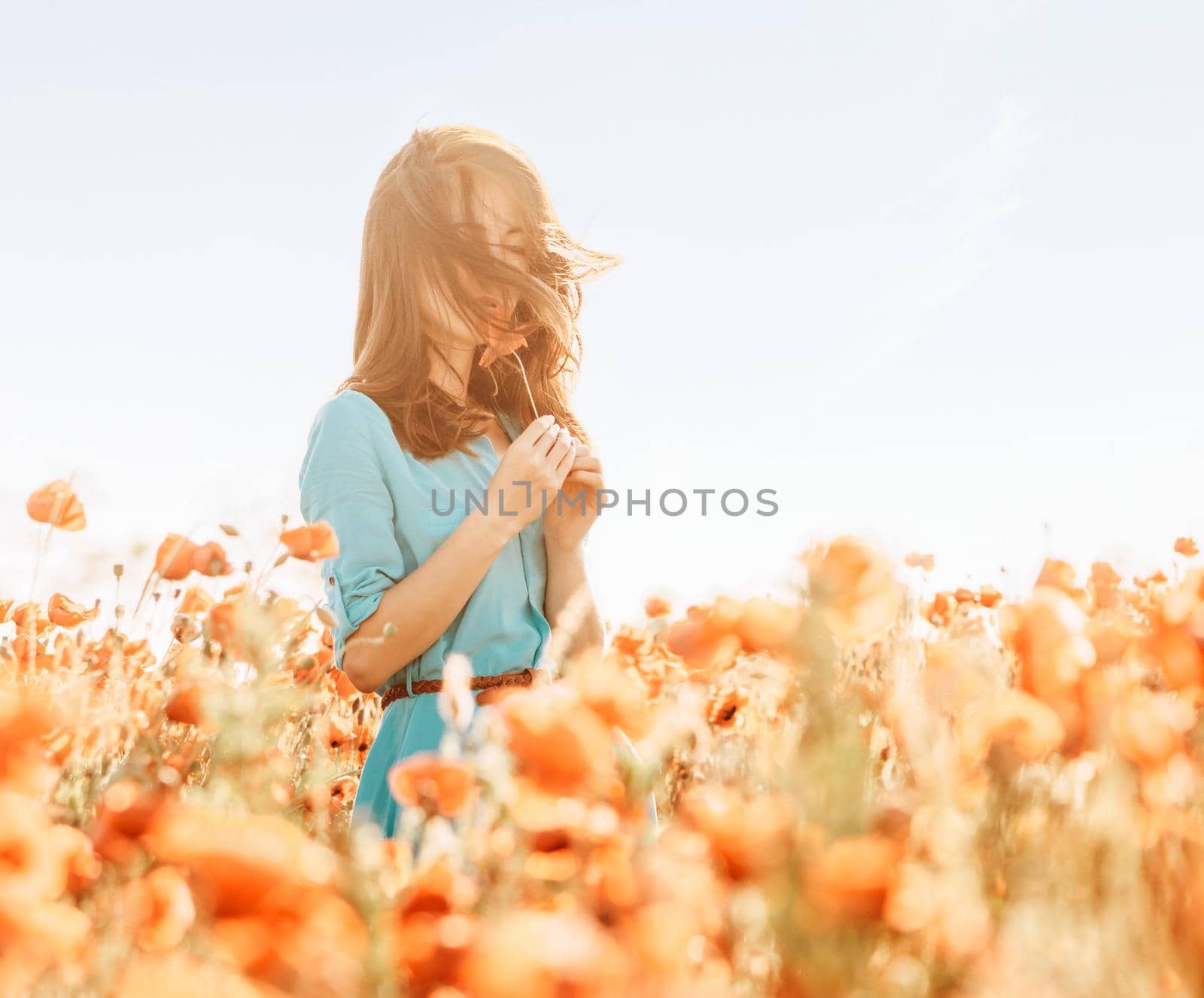 Beautiful young woman standing in poppies meadow and sniffing a flower in summer outdoor.