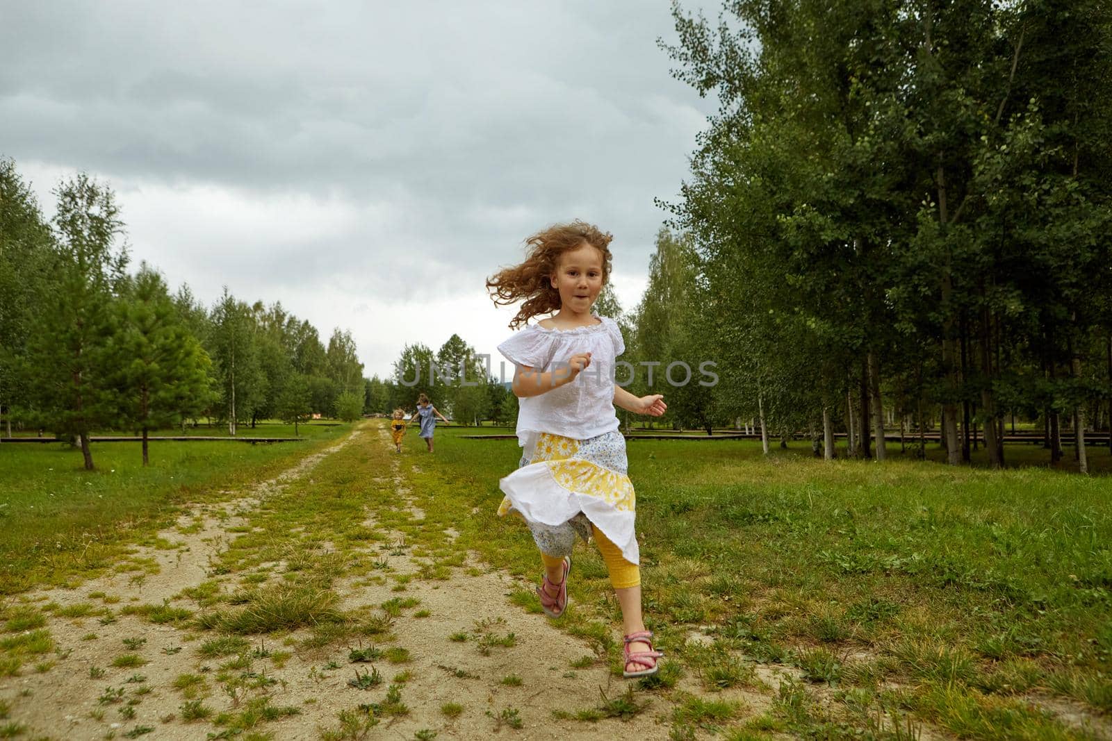 Funny girl in skirt and blouse running on road towards camera against cloudy sky while spending weekend day near grove in nature