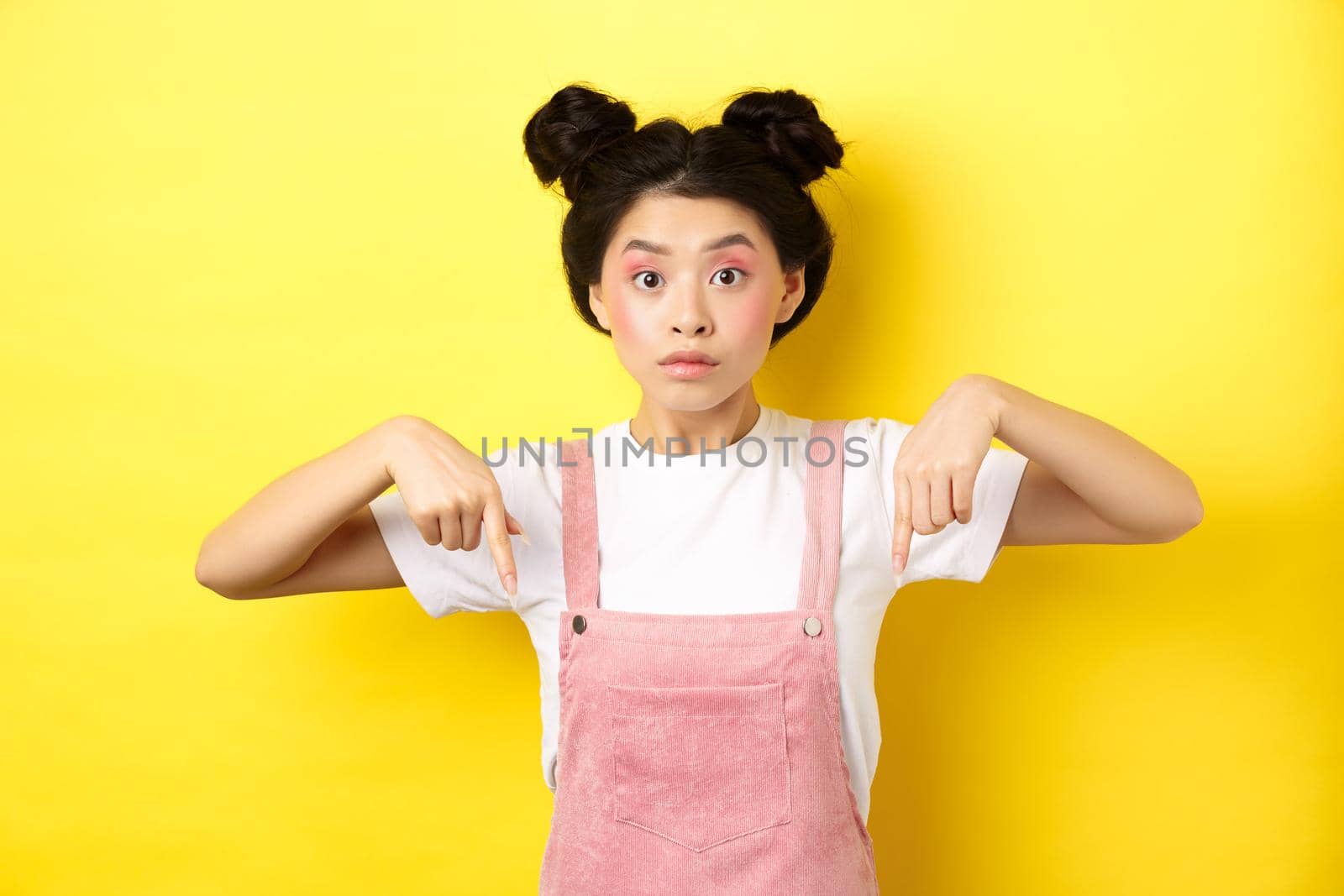 Surprised asian woman with beauty makeup, pointing fingers down and looking excited, showing interesting news, yellow background by Benzoix