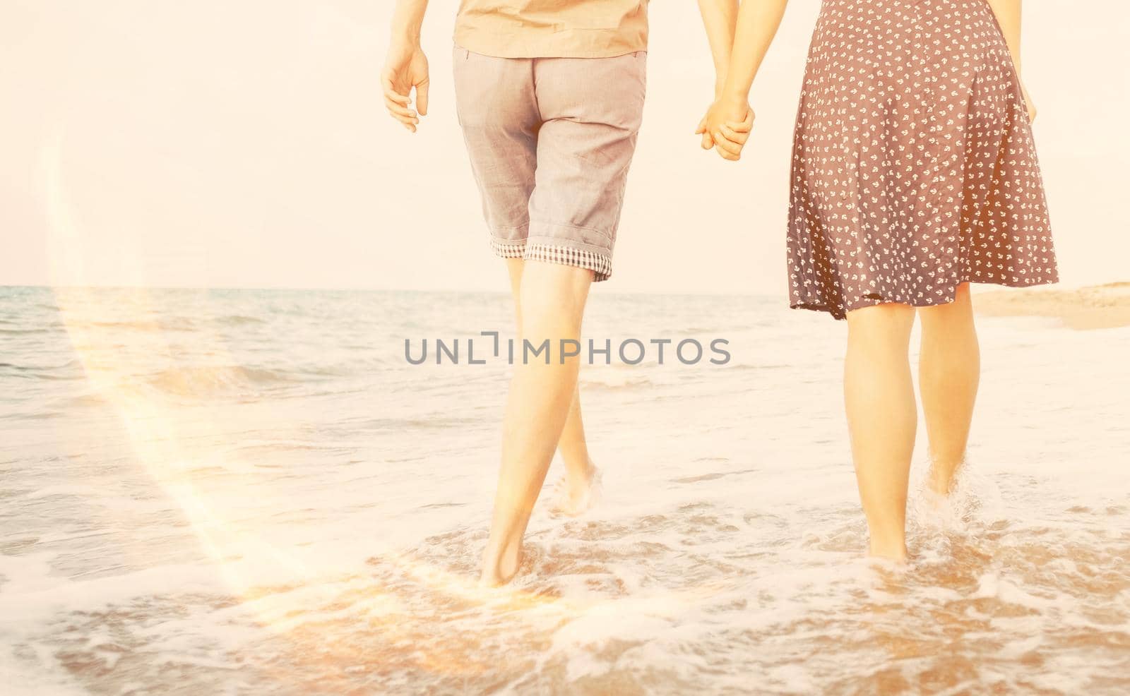 Unrecognizable young loving couple walking on sea coast and holding hands, rear view. Image with sunlight effect.