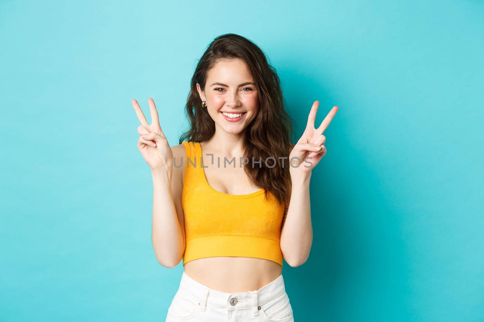 Summer holidays and emotions concept. Cute coquettish girl smiling and laughing, showing peace v-signs on blue background.