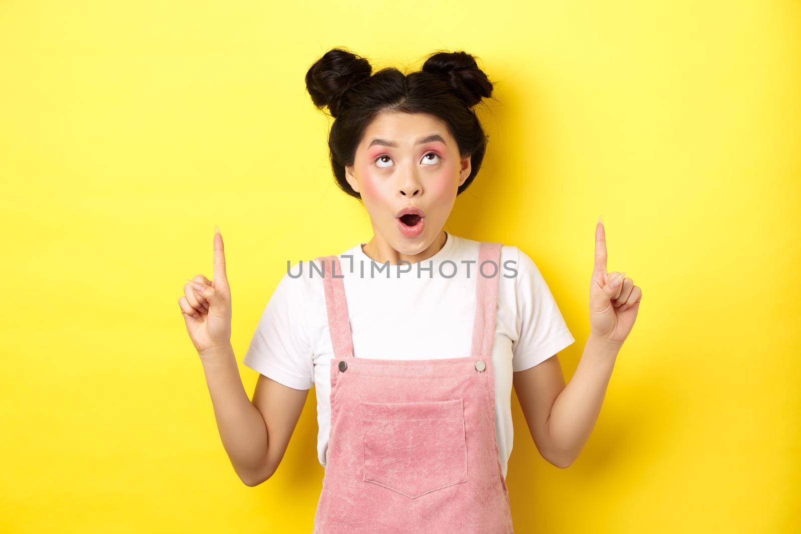 Excited asian teen student with glamour makeup, gasping amazed pointing fingers up, showing advertisement, yellow background by Benzoix