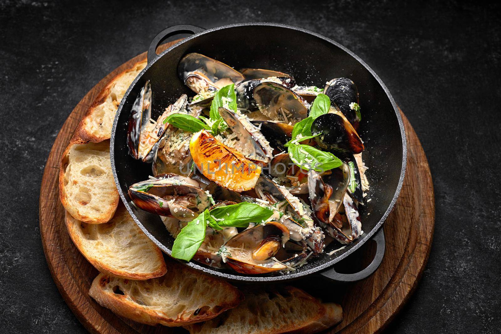 Cooked mussels in a skillet with cheese and basil leaves, on a wooden board, on a dark background