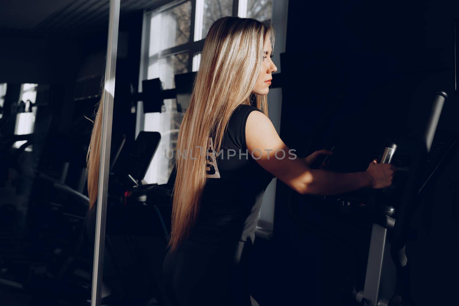 Attractive young blonde woman exercising on cardio training apparatus in dark gym