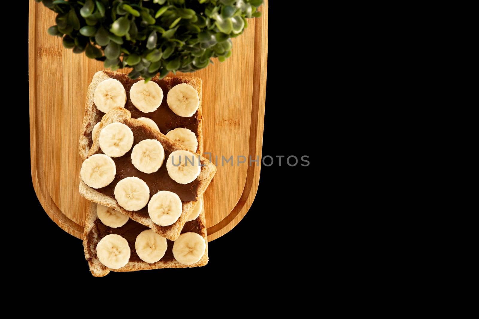 Three banana white bread toasts spread with chocolate butter that lie on a chopping board with a sprig of leaves on a dark background. top view with area for text