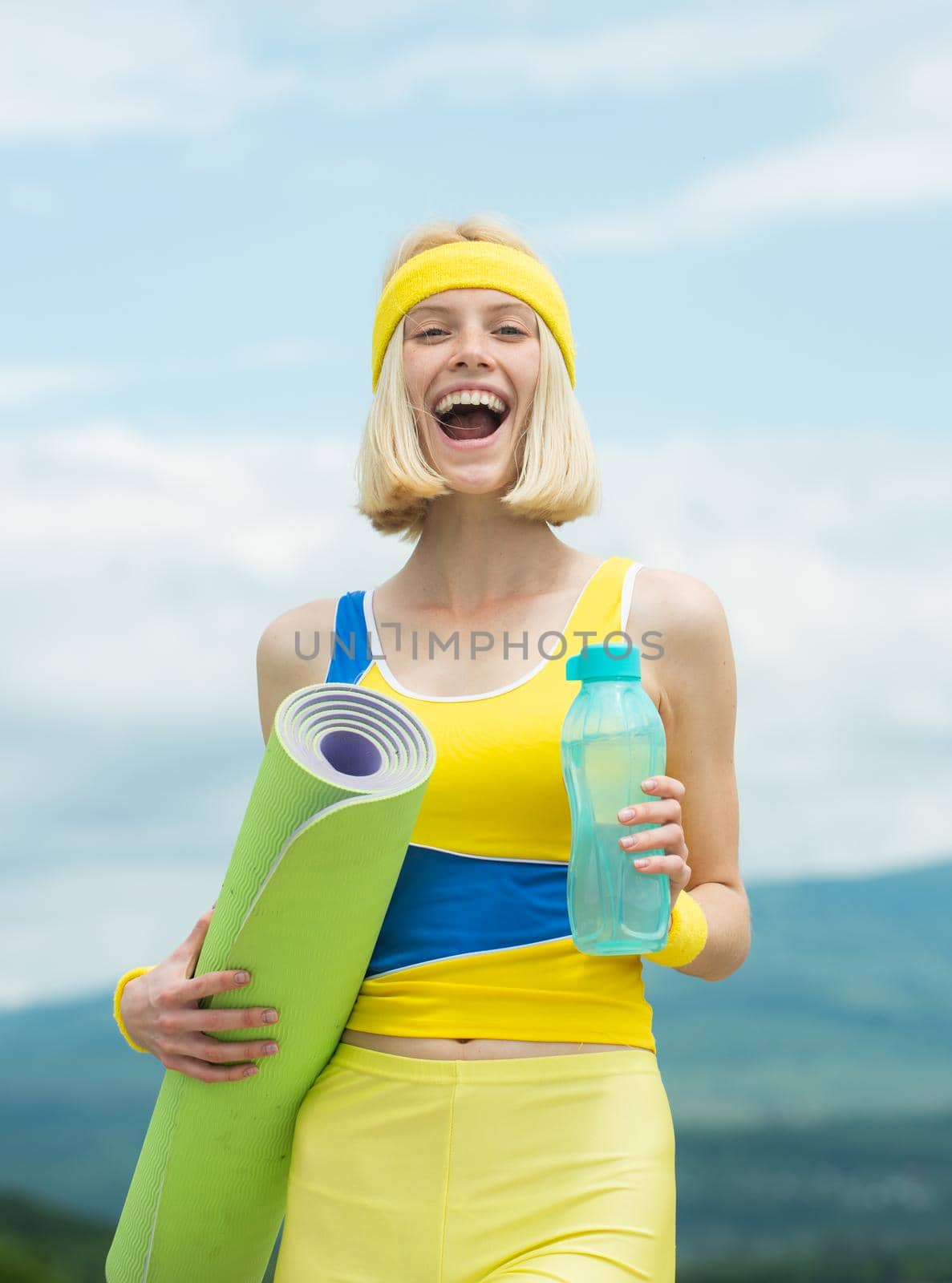 Excited amazed fitness trainer female waiting at you holding bottle of water and fitness mat, looking brave into camera while, wearing yellow-blue clothes training outside. by Tverdokhlib