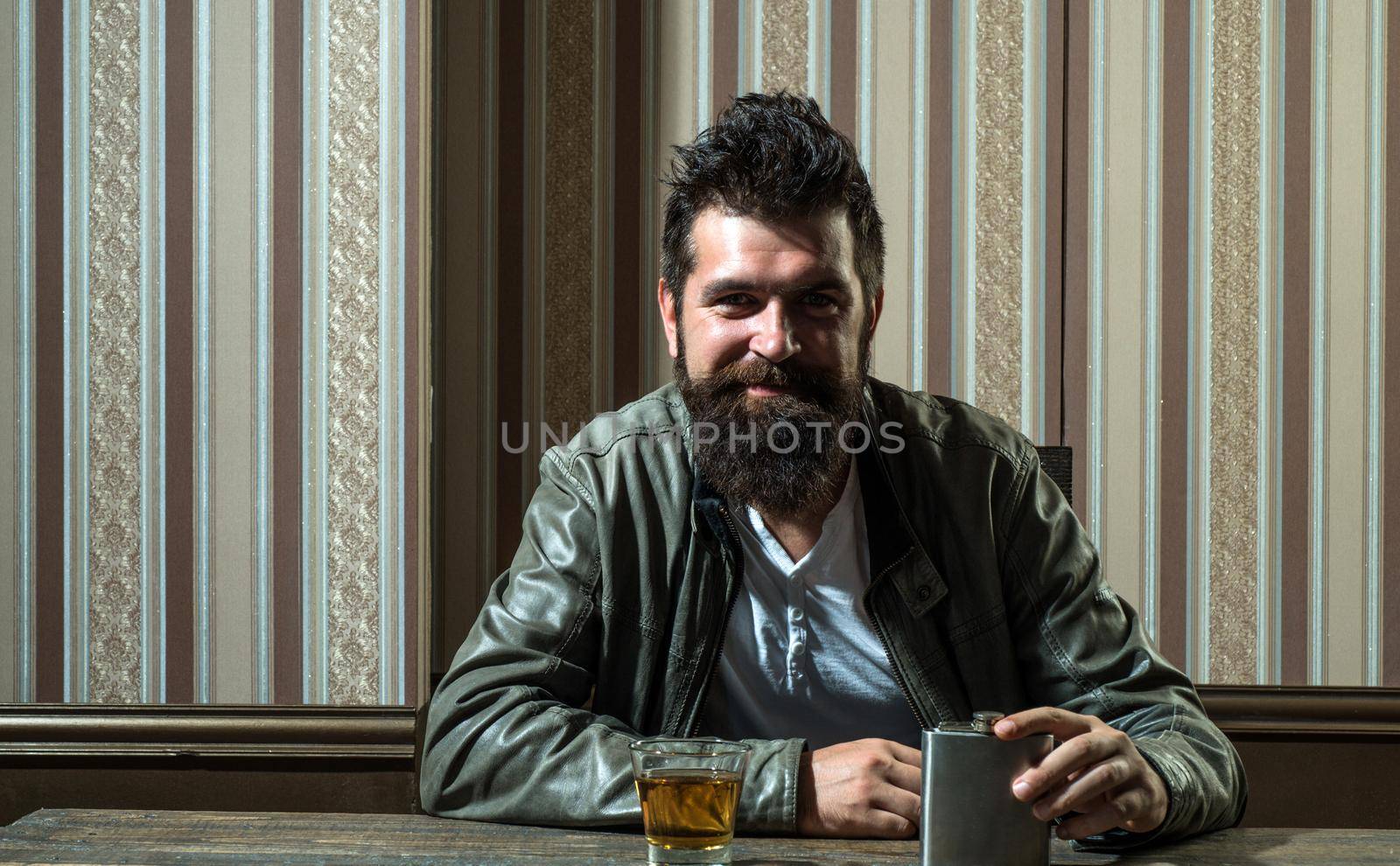Handsome smiling man drinking alone. Bearded brutal hipster man drinking expensive cognac. Serious problems with alcohol addiction. Alcoholism treatment. by Tverdokhlib