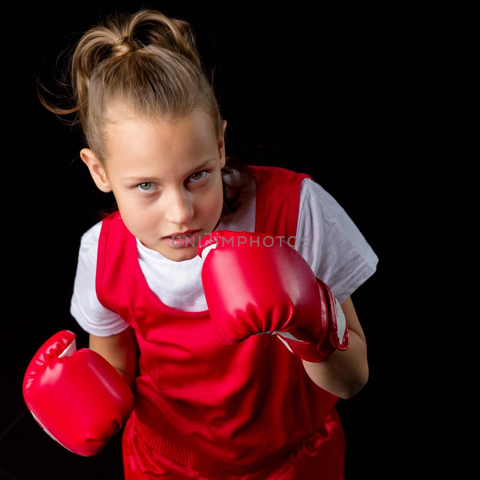 Sporty teenage girl doing boxing exercises. Close up portrait of pretty girl in red sports uniform making direct hit. Preteen child posing in studio against black background