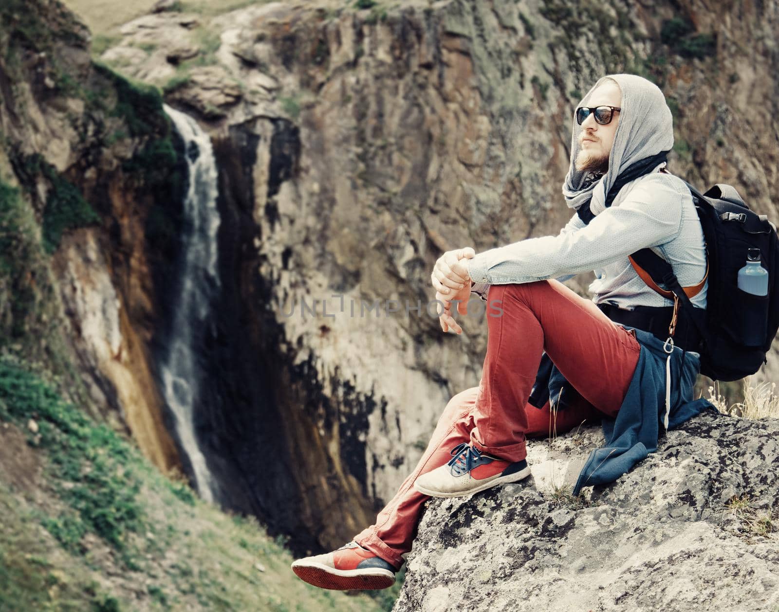 Traveler young man sitting on cliff on background of waterfall outdoor.