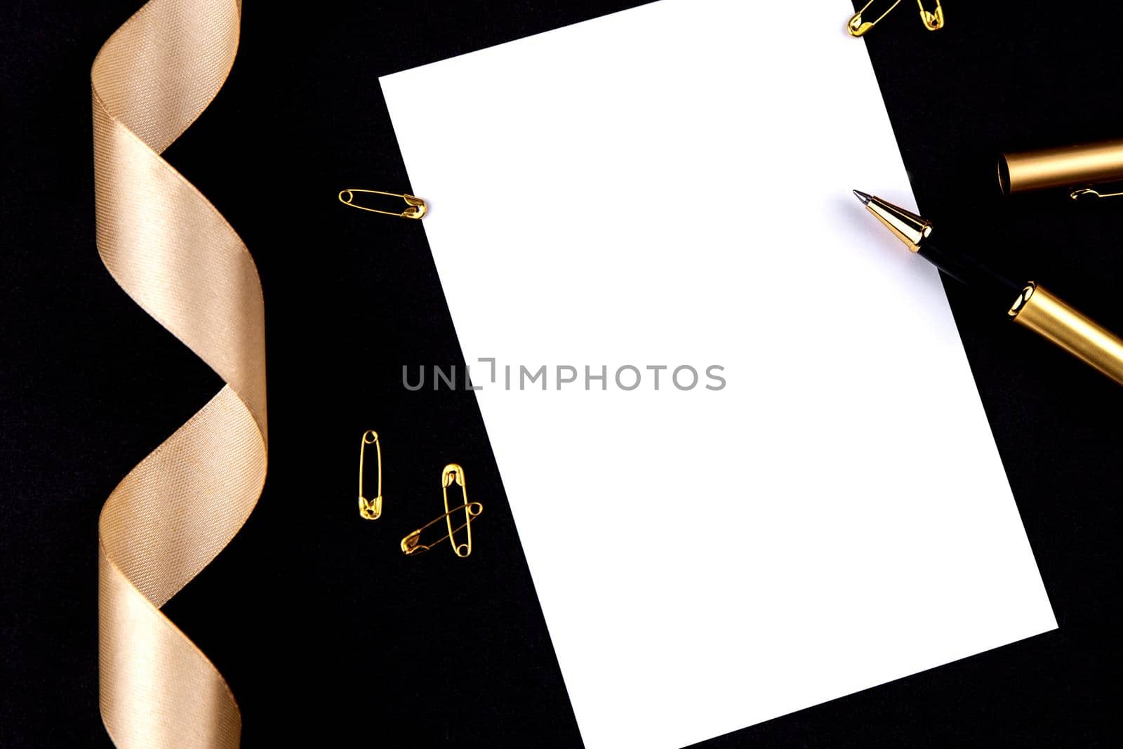 Gold pen, ribbon, paper clips and stationery on a black background with a white sheet of paper.