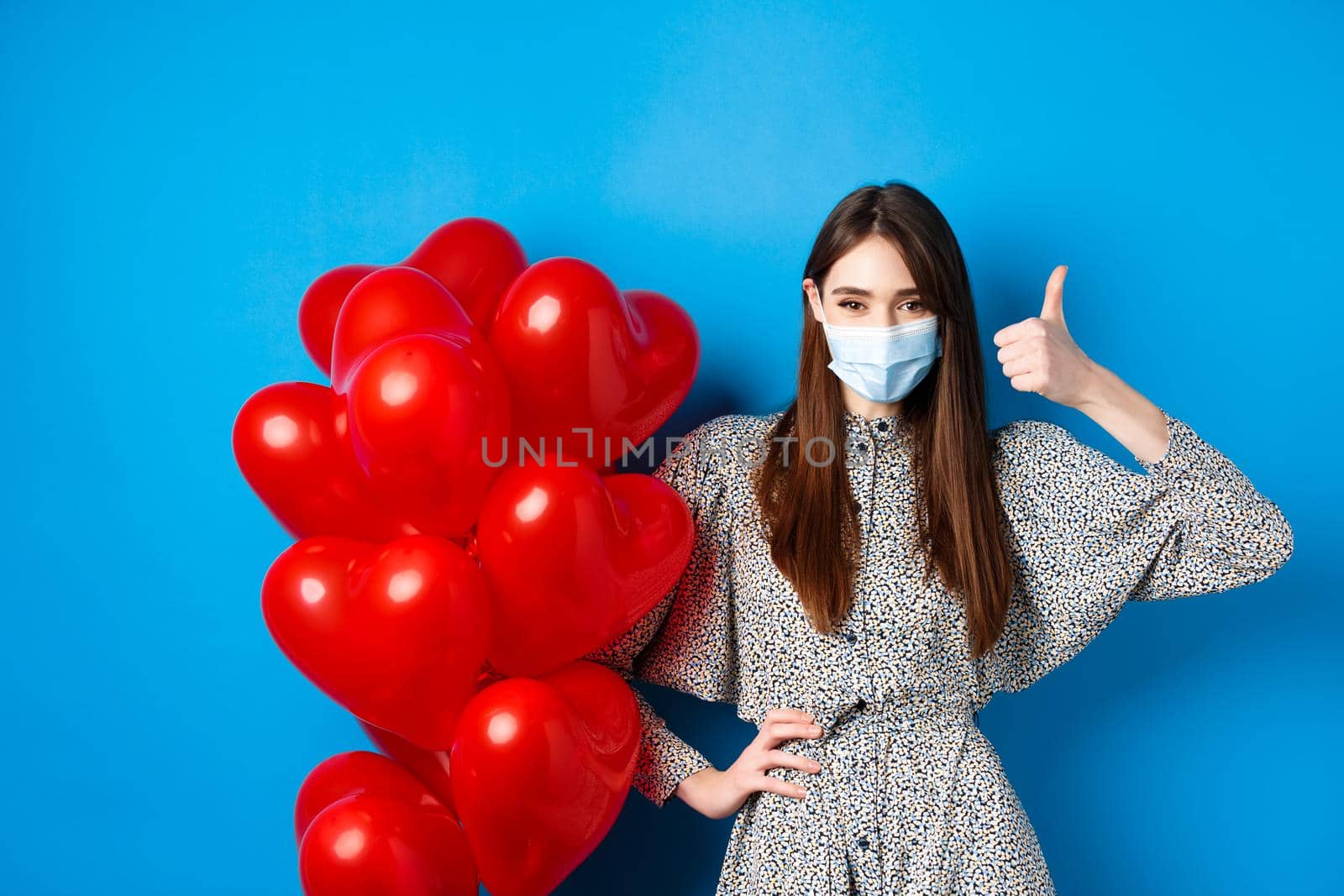 Coronavirus and pandemic concept. Beautiful woman in medical mask and dress standing near Valentines day balloons and showing thumb up, standing on blue background.