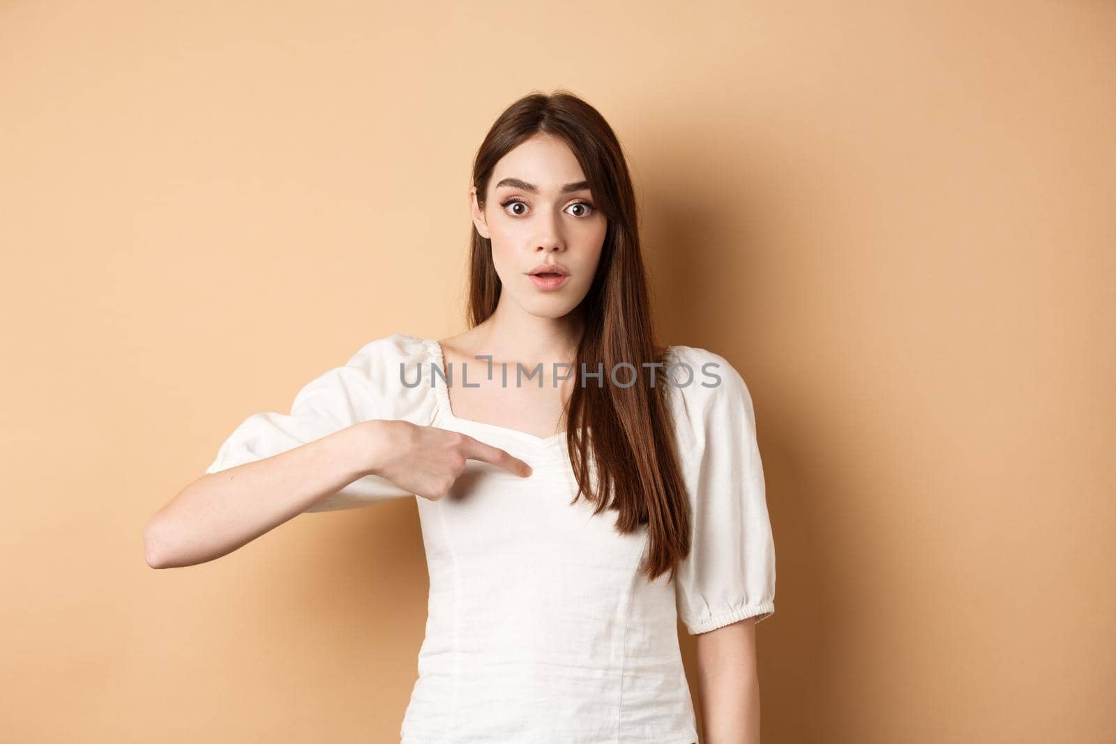 Confused girl pointing at herself with disbelief, being chosen or picked, standing on beige background by Benzoix