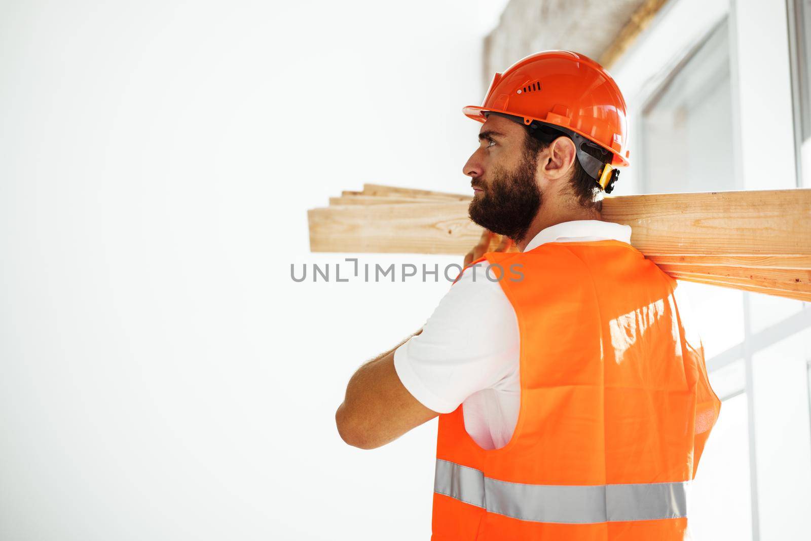 Builder man in hardhat carrying timber on building site, close up . by Fabrikasimf