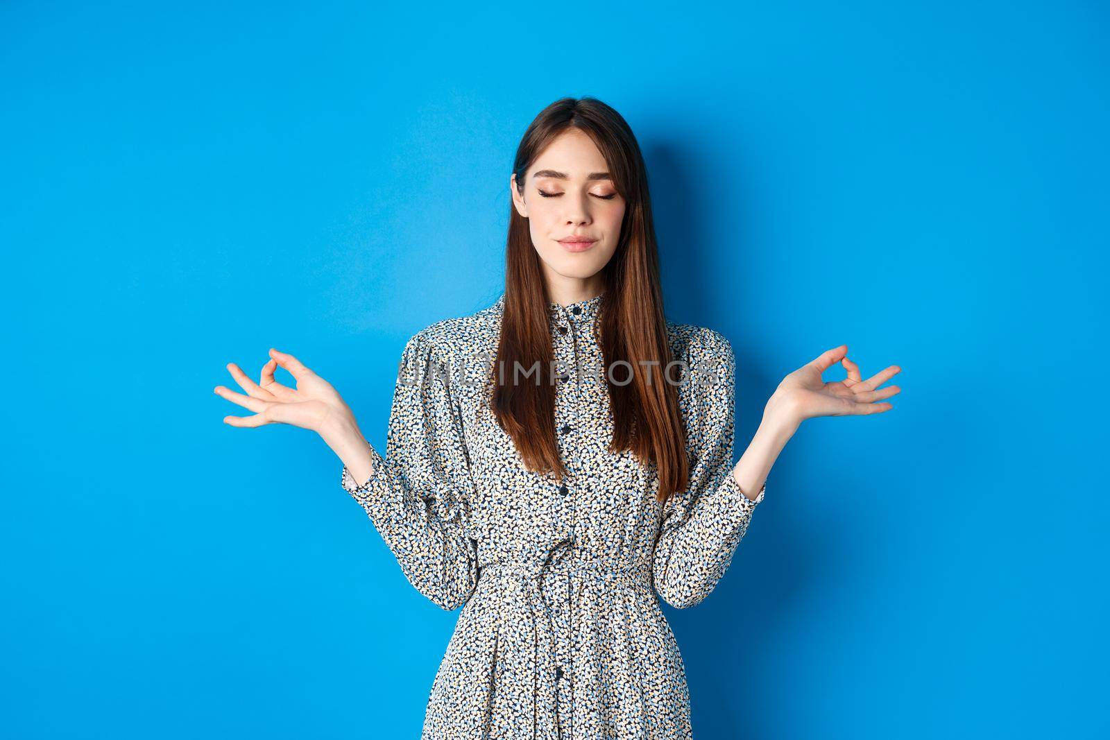 Mindful young woman in dress, close eyes and smile, meditating peaceful, standing with hands spread sideways with zen sign, blue background.