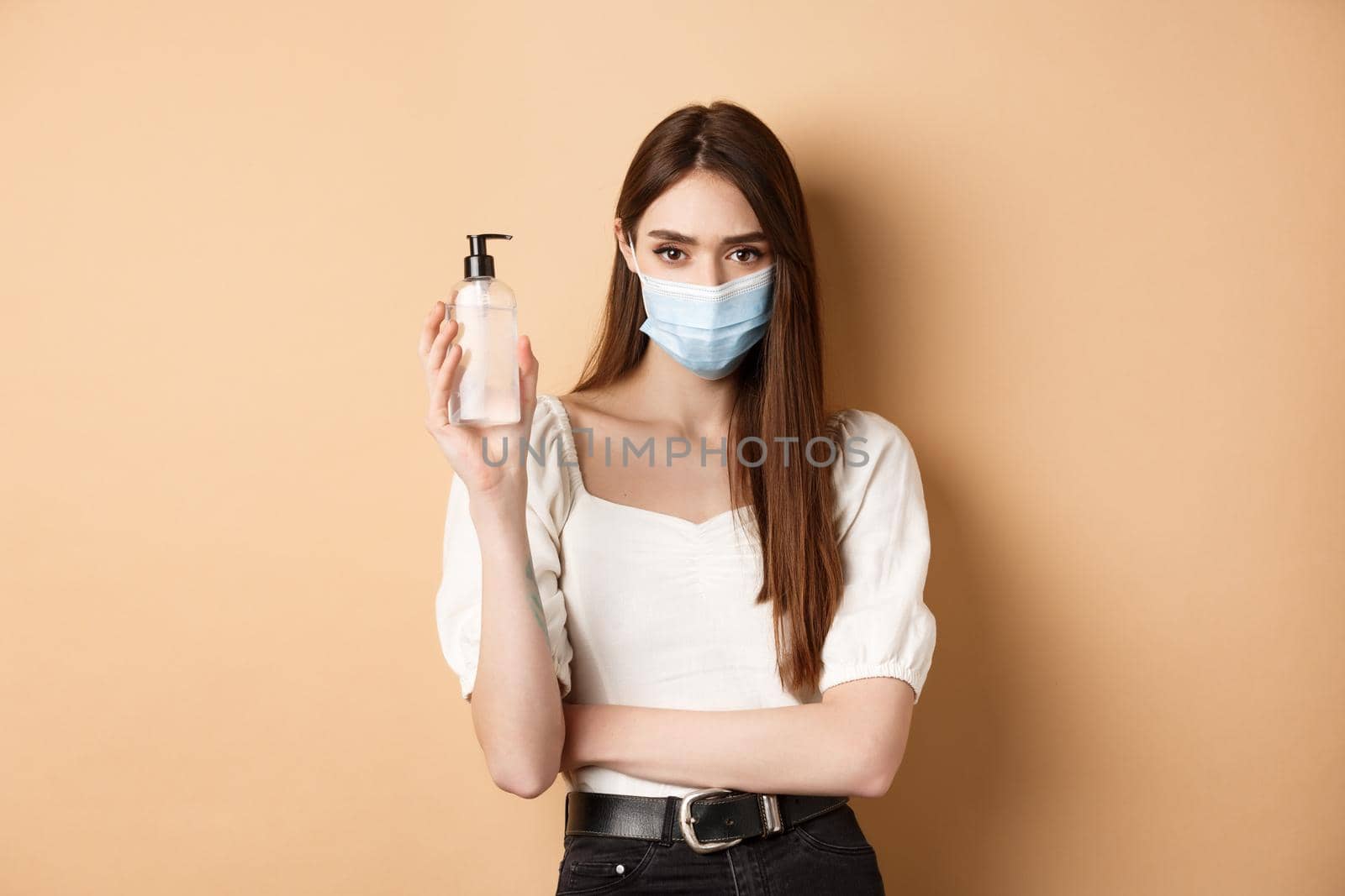 Pandemic and healthcare concept. Serious woman concerned about covid-19, wearing medical mask and showing bottle of hand sanitizer, standing on beige background.