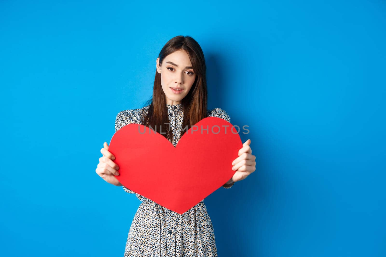 Valentines day and relationship concept. Tender young woman in dress stretch out hand and giving big red heart to you, making confession, standing romantic on blue background by Benzoix