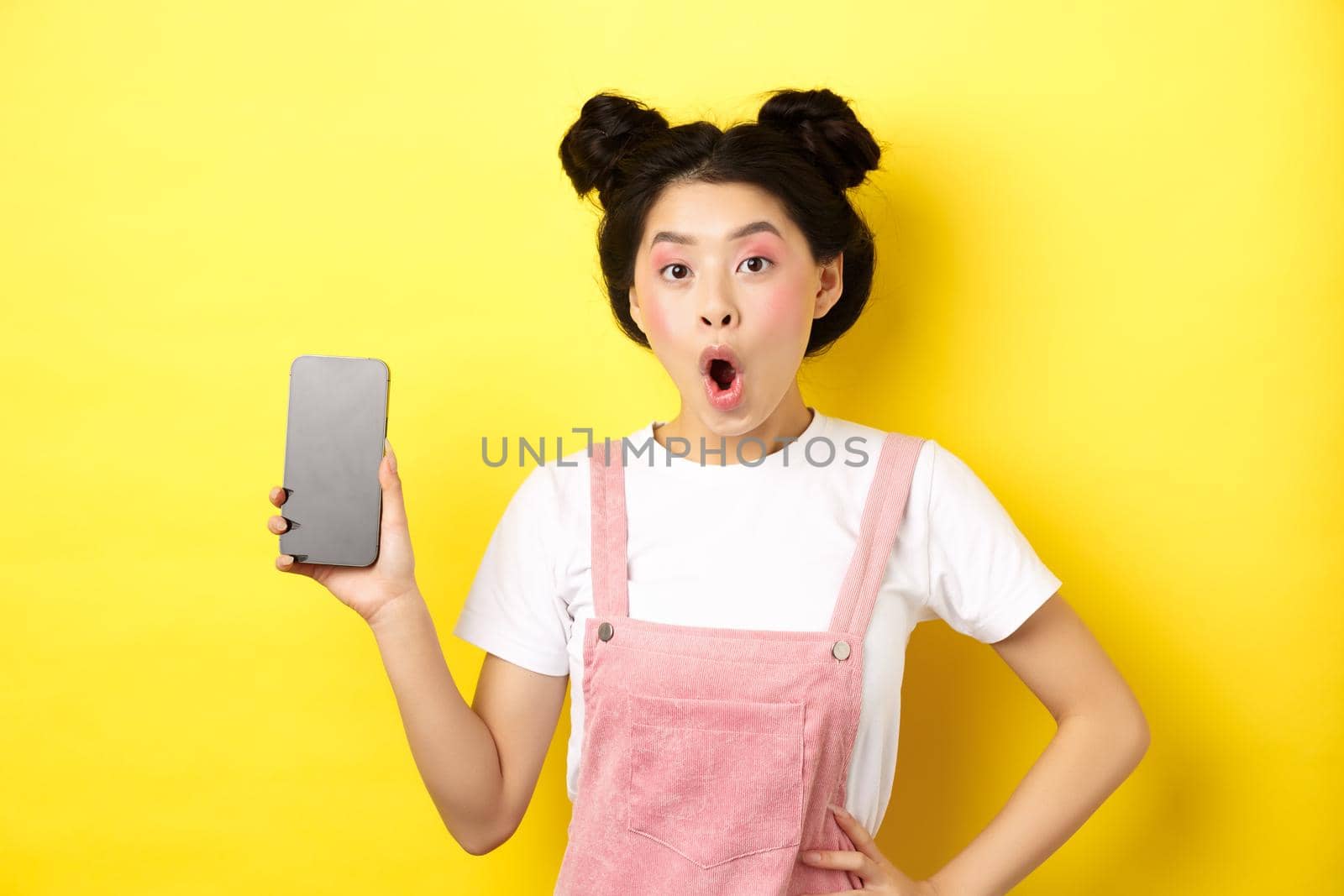 Online shopping concept. Excited stylish asian girl say wow, showing empty cell phone screen and look amazed, yellow background by Benzoix