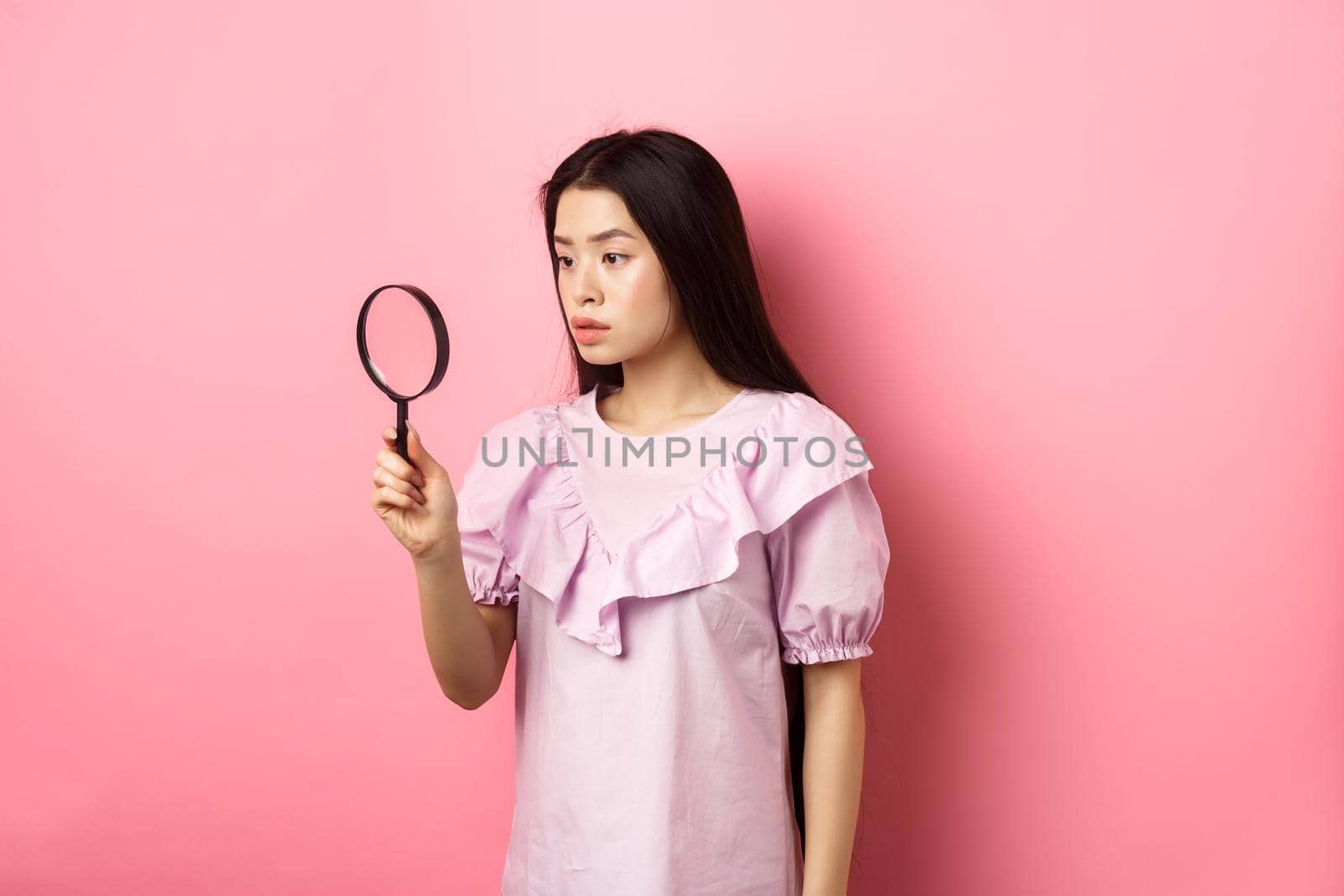 Serious girl investigating, looking through magnifying glass, searching for something, standing against pink background.