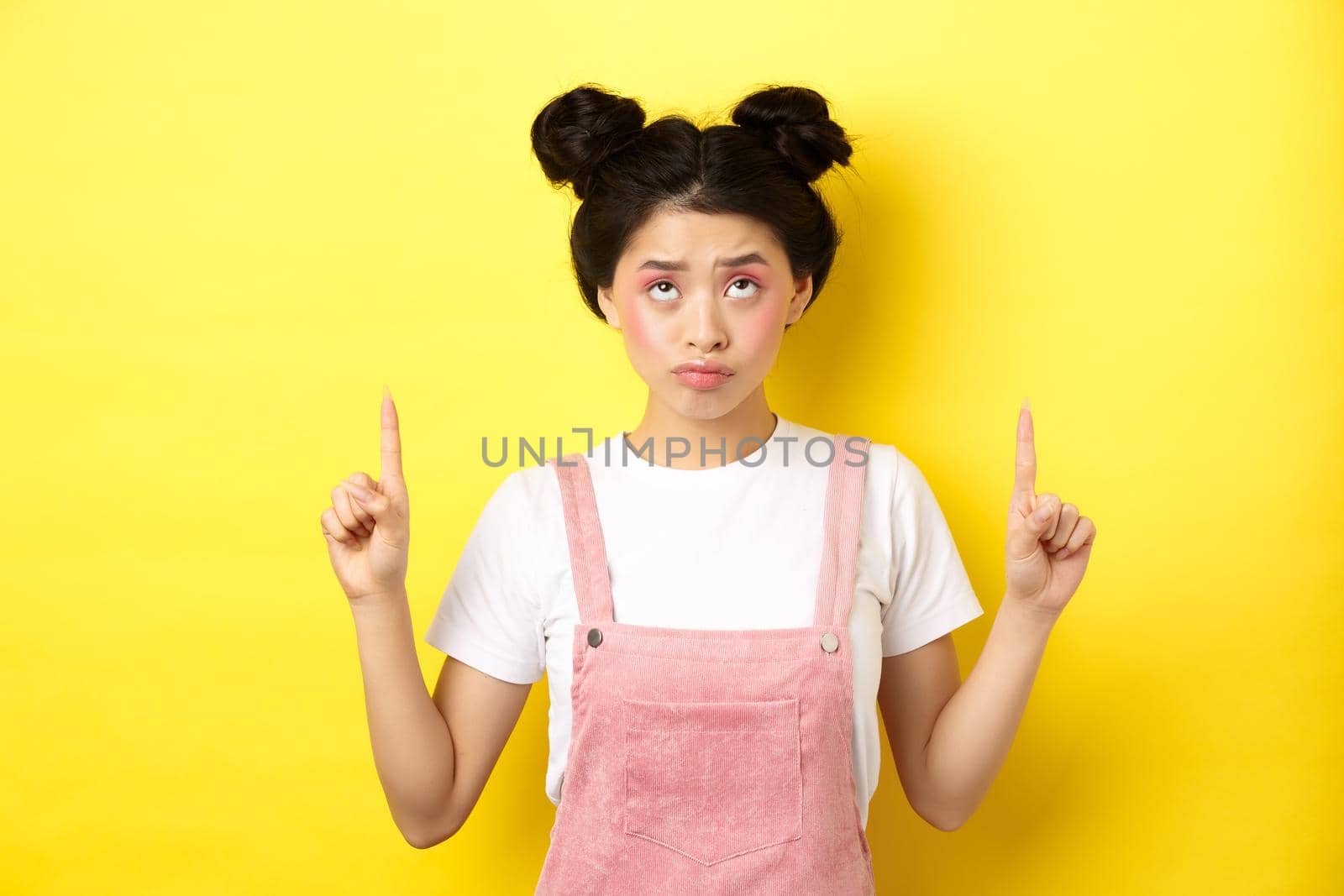 Skeptical teenage asian girl with glam pink makeup, pointing and looking up unamused, standing reluctant on yellow background.