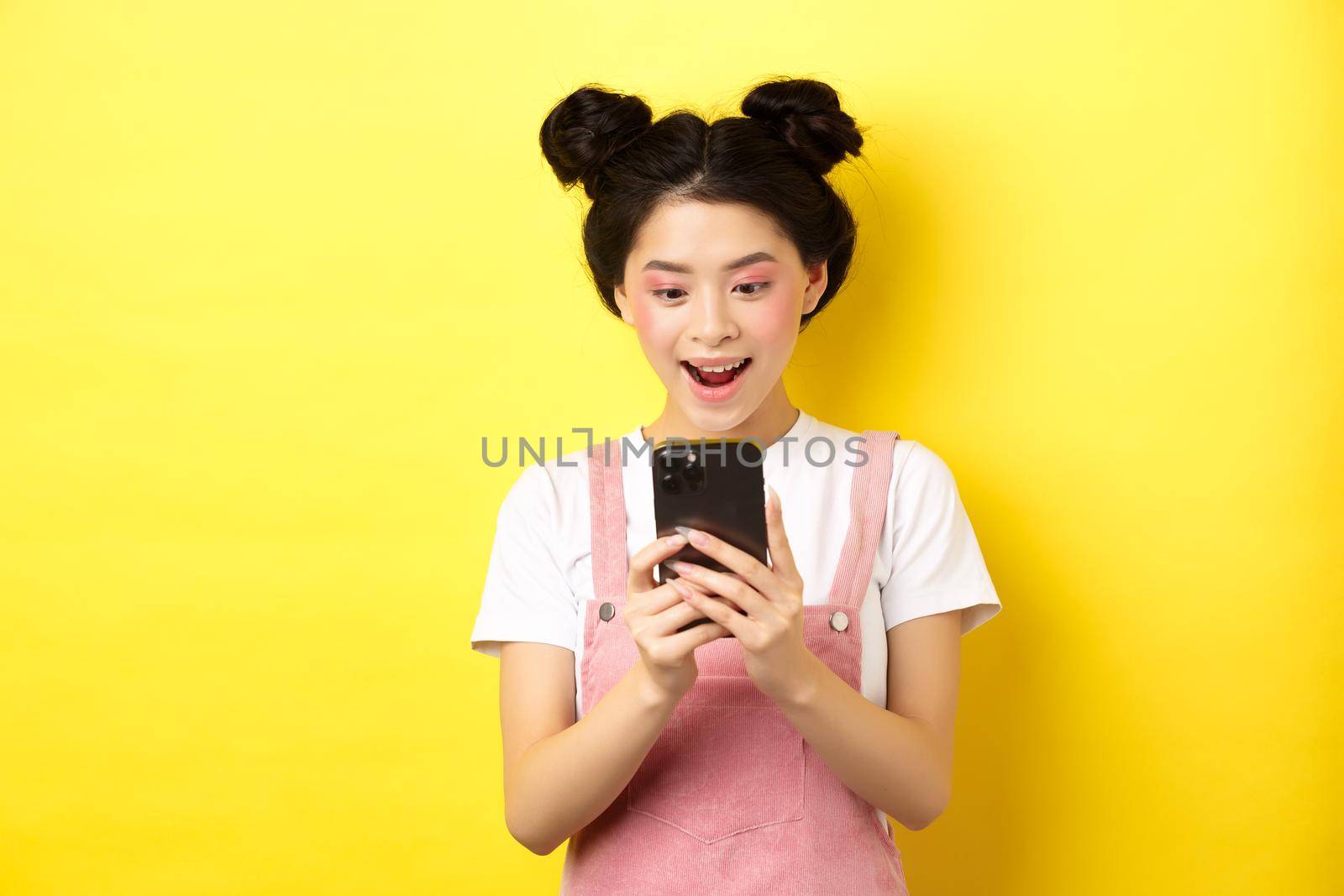 Pretty asian girl looking excited at screen, reading message on phone and smiling happy, standing in summer clothes on yellow background.