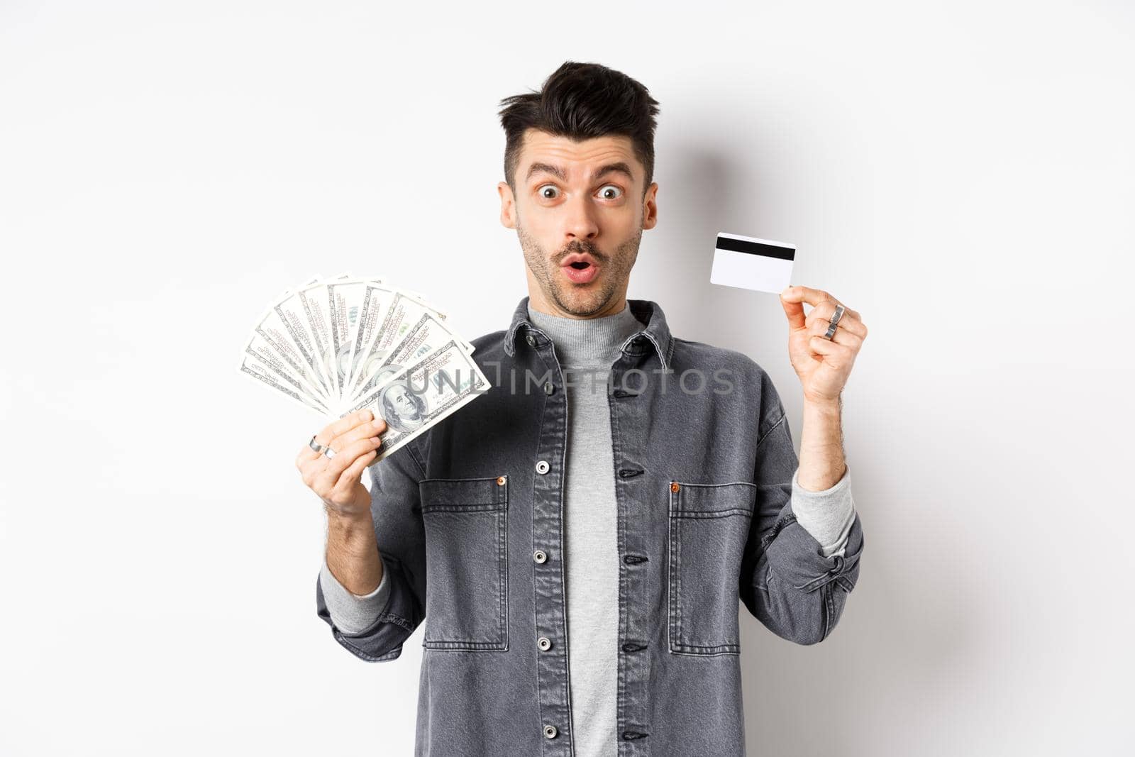 Excited guy holding plastic credit card and dollar bills, standing amused on white background by Benzoix