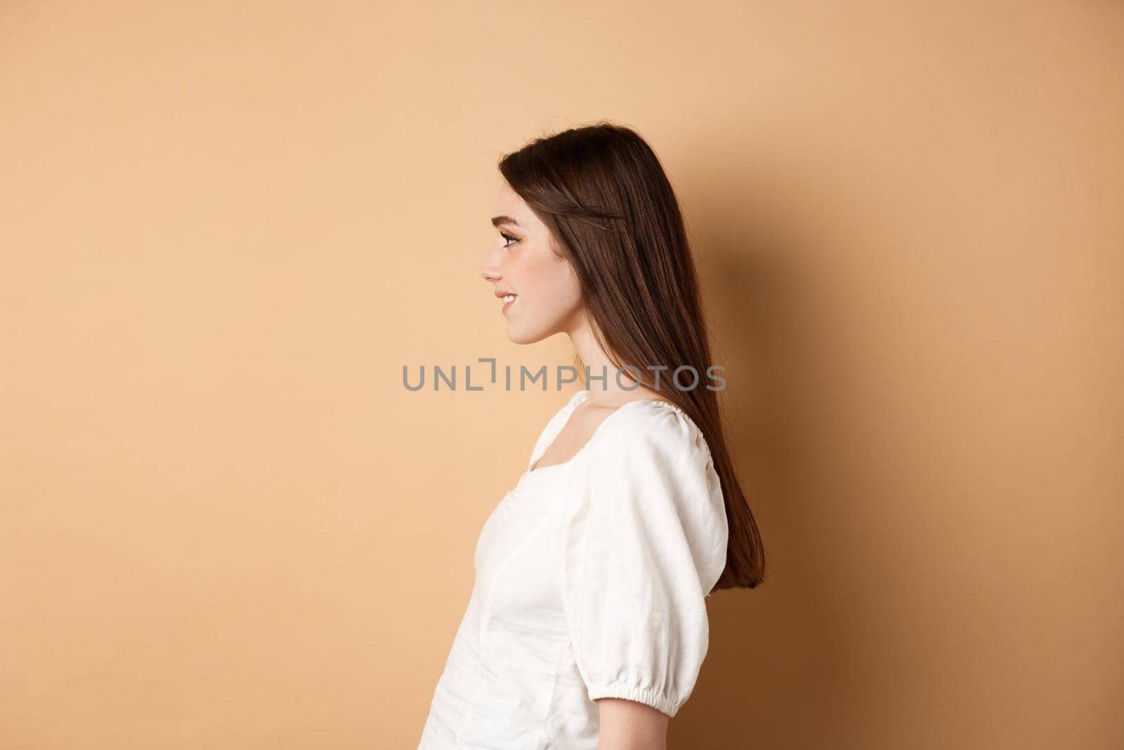 Profile of beautiful caucasian woman with long natural hair, looking left at empty space and smiling romantic, standing on beige background.