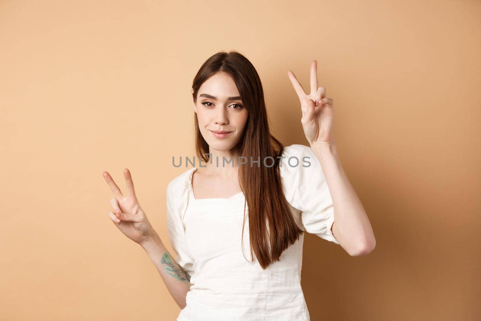 Cheerful attractive girl dancing and having fun, showing v-signs and looking positive at camera, standing on beige background.