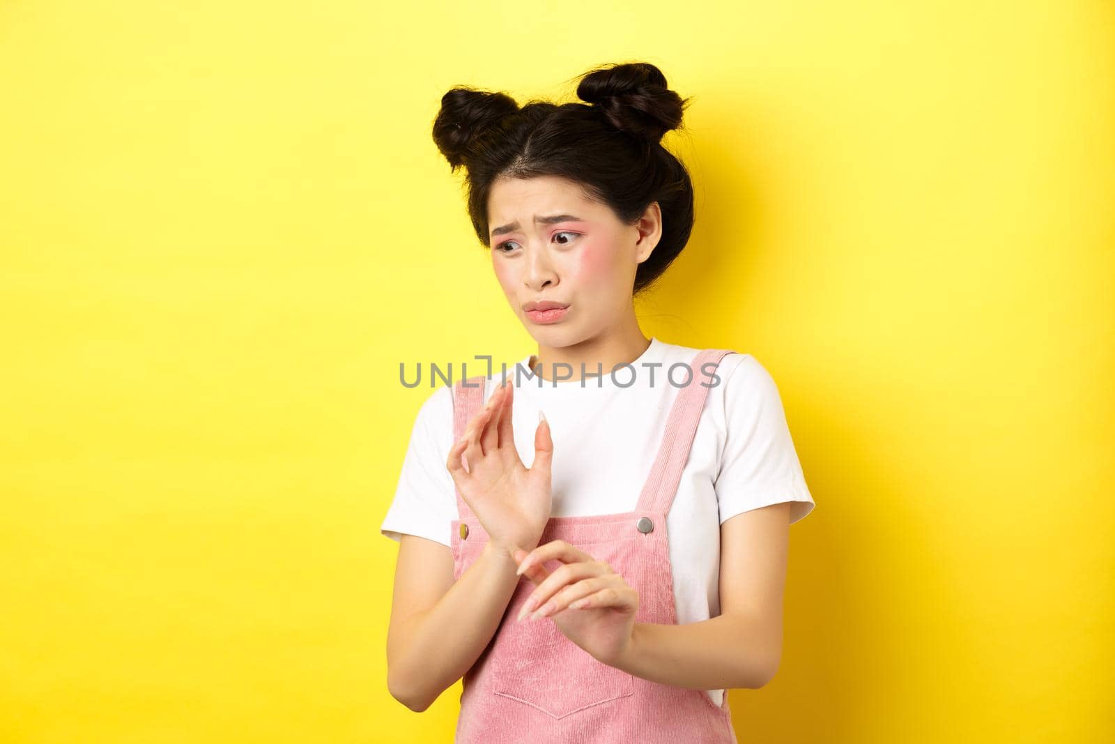 Disgusted asian girl raising hands up defensive, block something disgusting, turn away from aversion and reluctance, standing on yellow background.