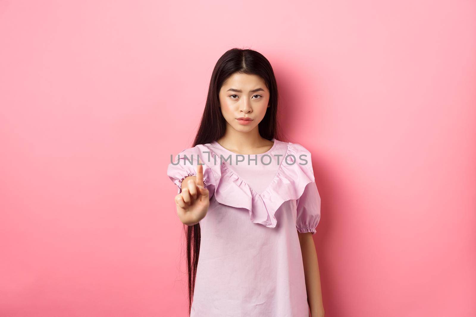 She says no. Serious asian girl shaking finger in stop gesture, prohibit and disagree with person, scolding bad behaviour, standing against pink background by Benzoix