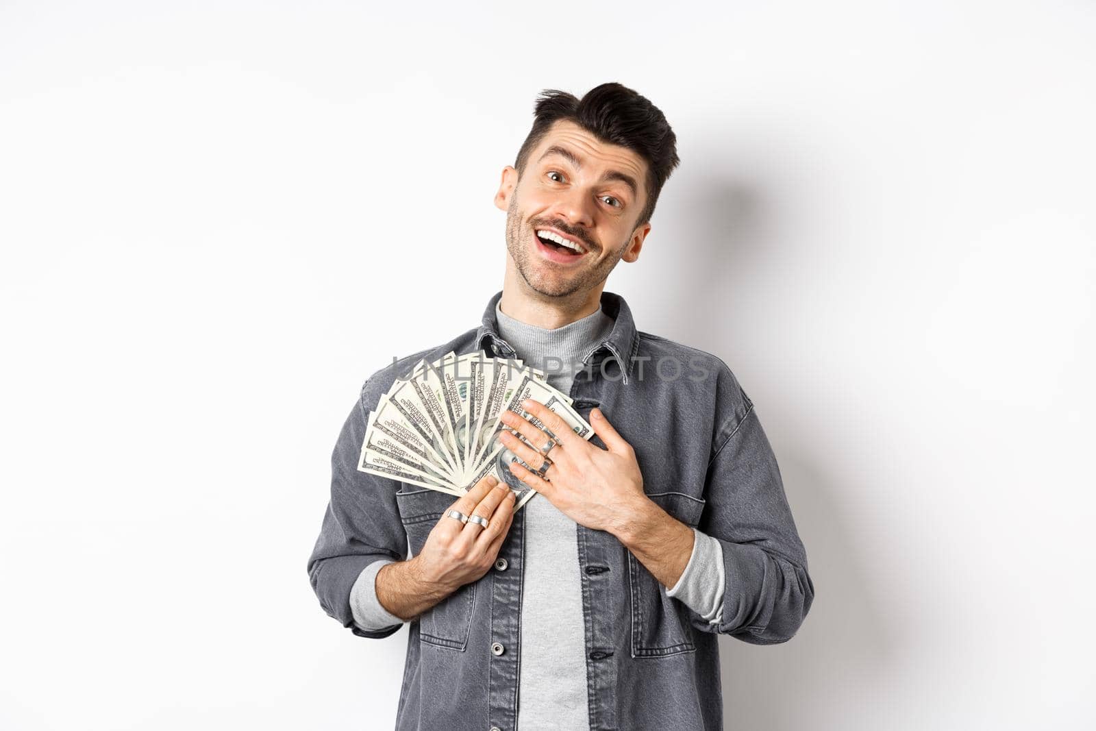 Happy smiling guy making money, hugging dollar bills with pleased relieved face, standing against white background.