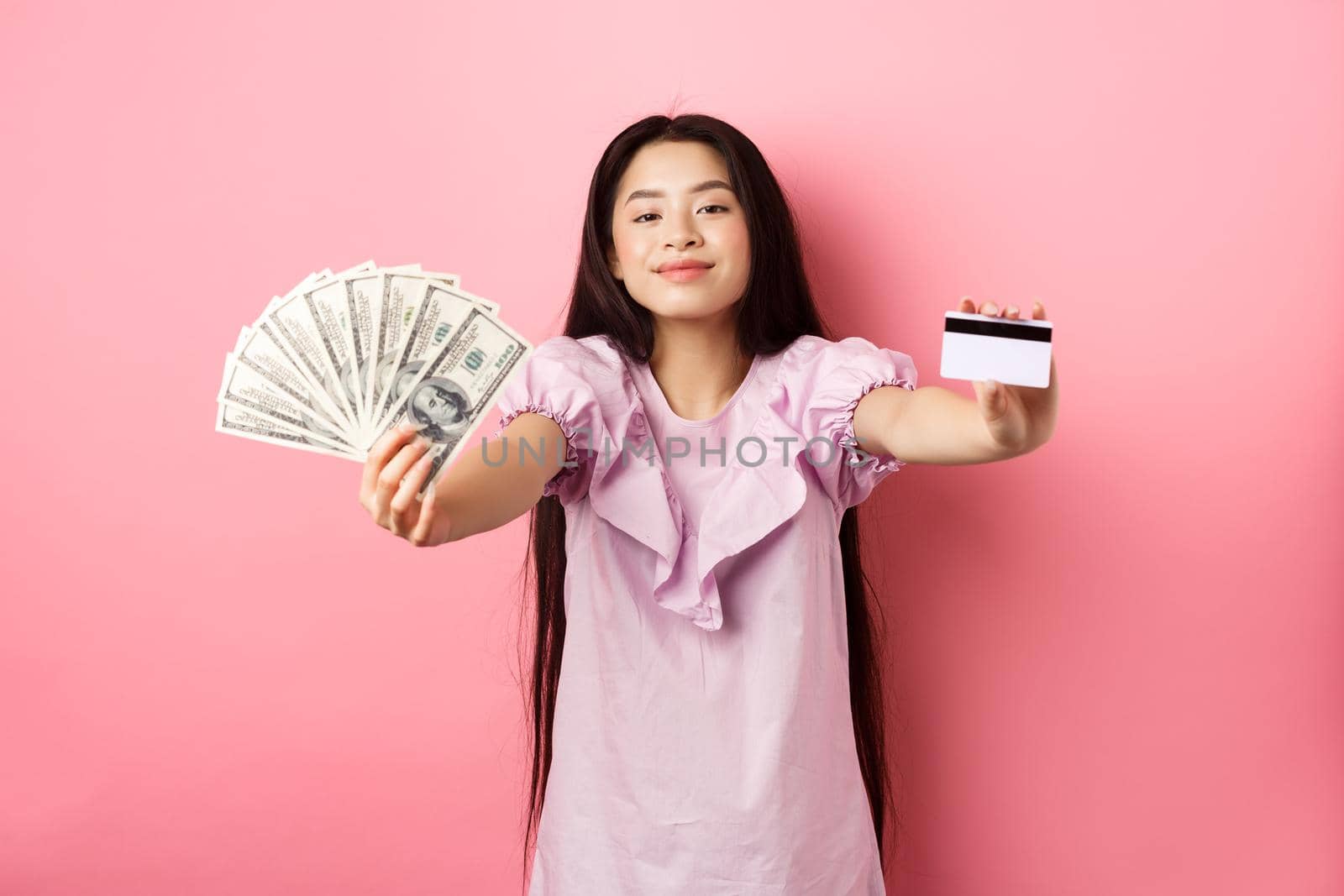 Beautiful asian woman showing plastic credit card and dollar bills, smiling pleased, standing against pink background.