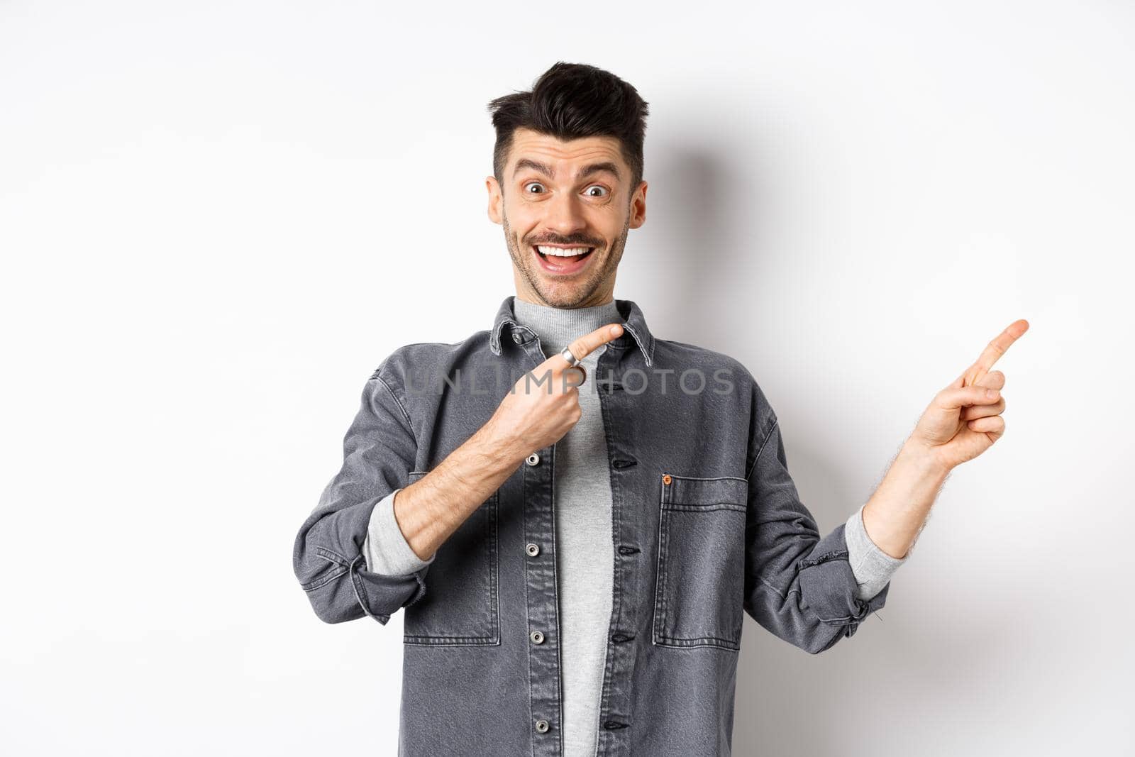 Look here. Happy stylish man smiling, pointing fingers right at empty space, showing cool promo offer, standing in denim jacket on white background.