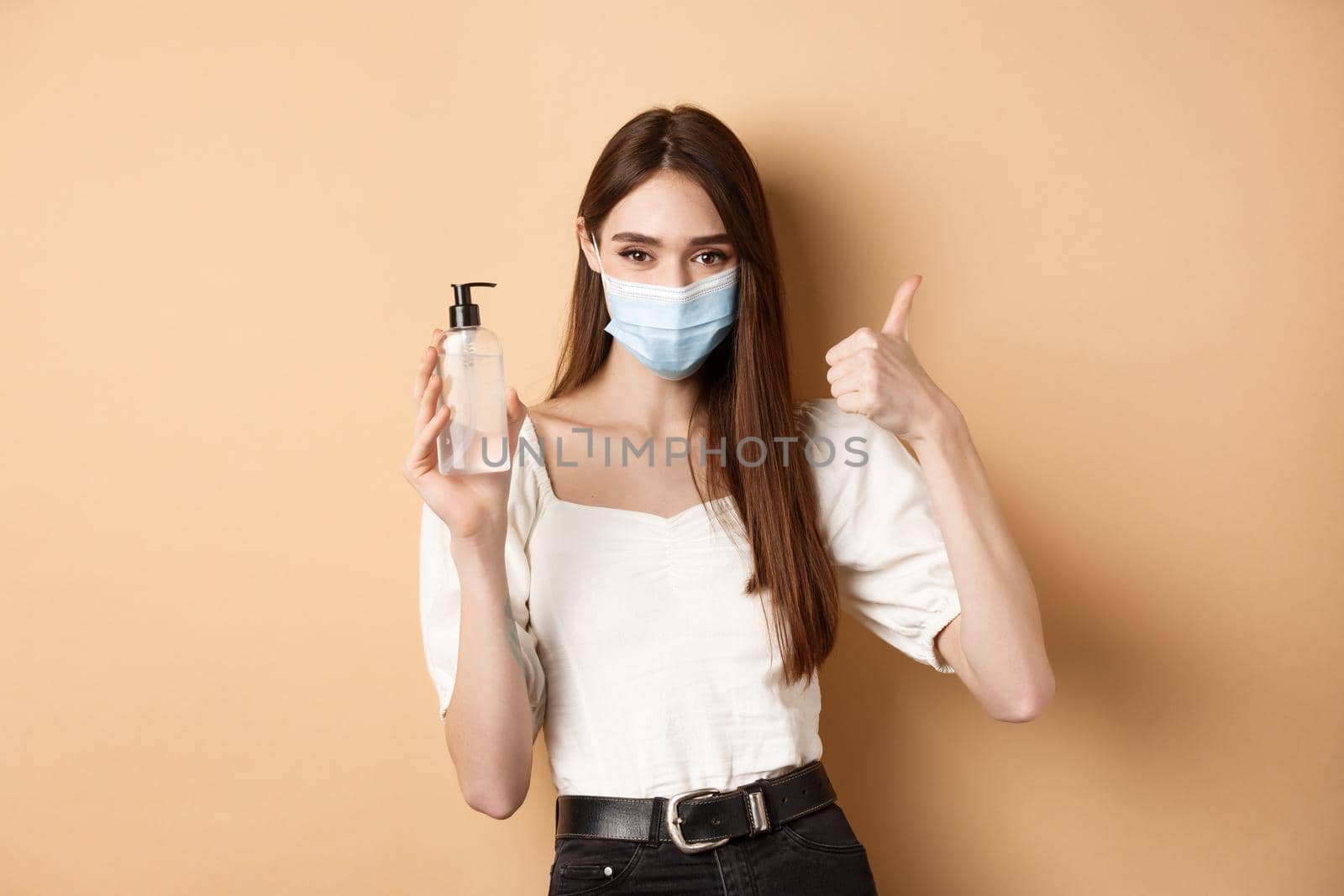 Covid-19 and preventive measures concept. Smiling girl in medical mask show thumb up and hand sanitizer, recommend product for disinfection, beige background.