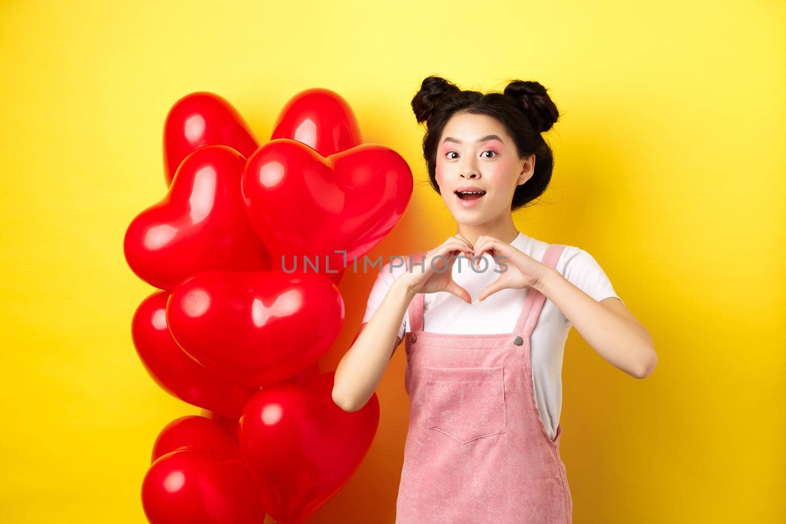 Happy asian girl showing heart sign near romantic balloons, waiting for true love on Valentines day, looking hopeful at camera, yellow background.