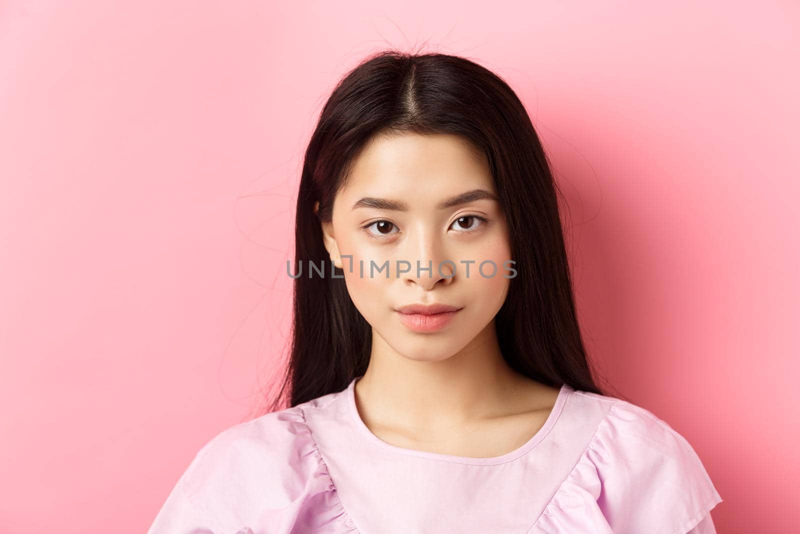 Close-up of beautiful young asian woman in dress, looking confident at camera, standing on pink background.