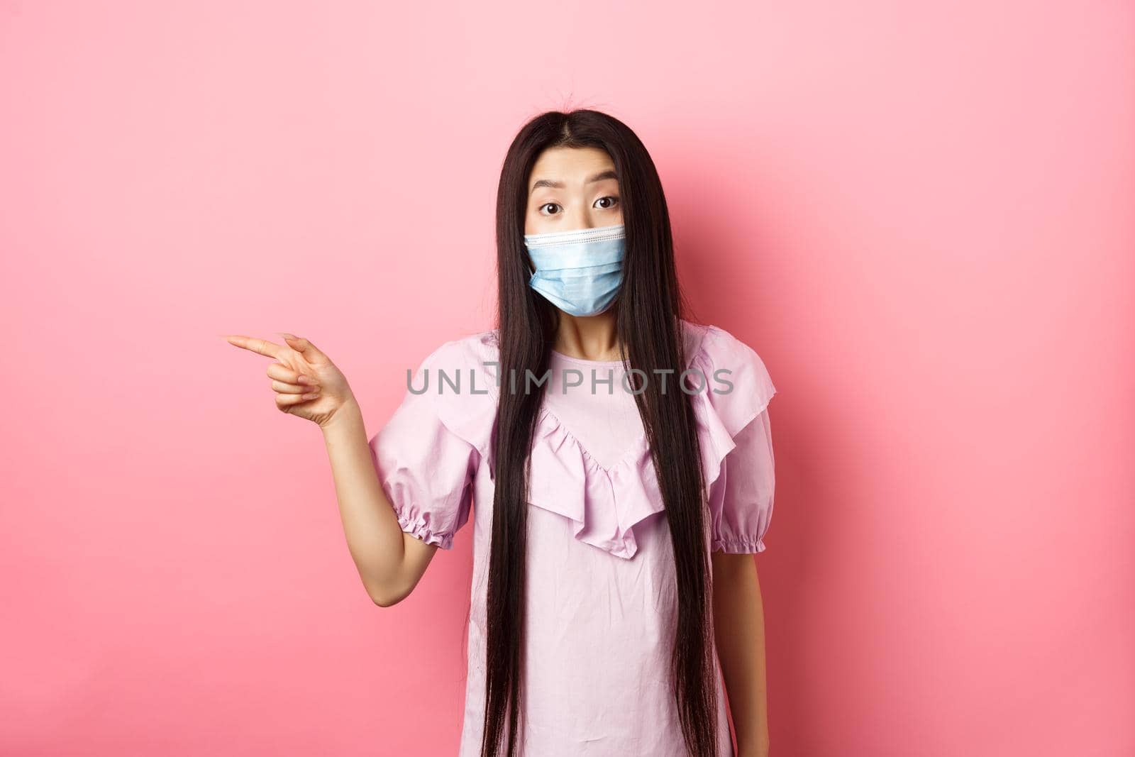 Covid-19, pandemic lifestyle concept. Curious asian girl in medical mask pointing left at logo, asking question about promotion, standing on pink background.