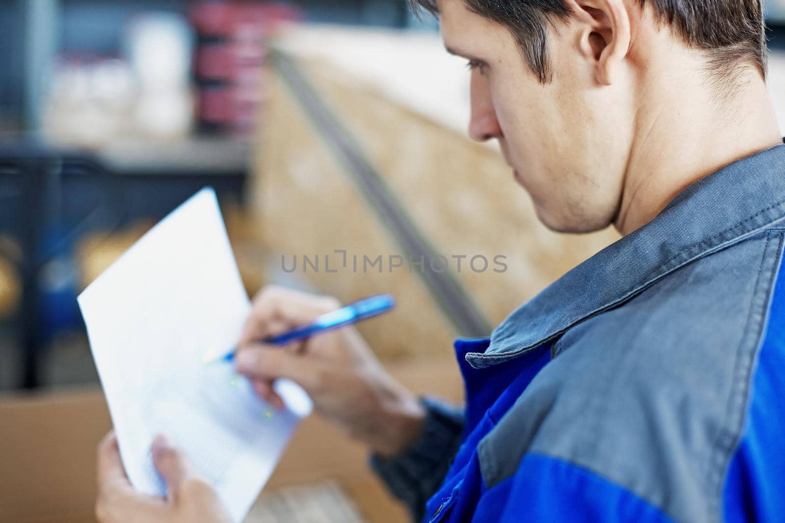 A warehouse worker in blue overalls accepts and describes the goods in the room. A young man of Caucasian appearance takes the goods in the warehouse and makes entries in the magazine. Attentive and focused employee.