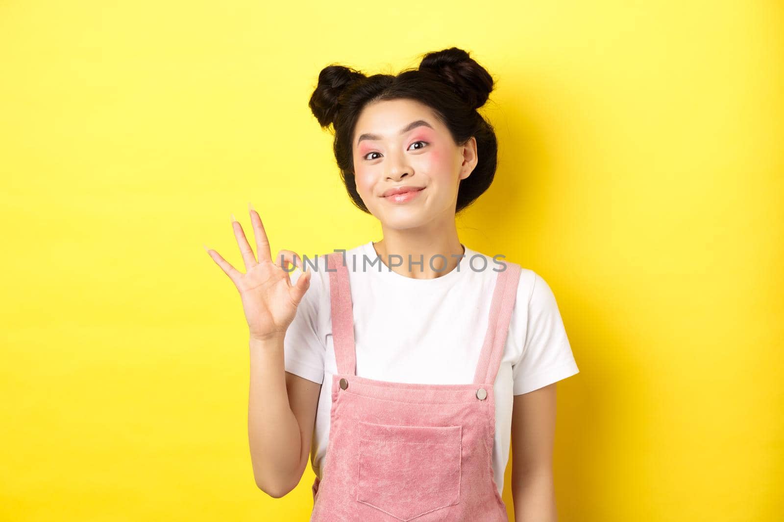 Cheerful asian beauty girl with makeup, show okay sign and smiling excited, approve good promo, recommending product, yellow background.