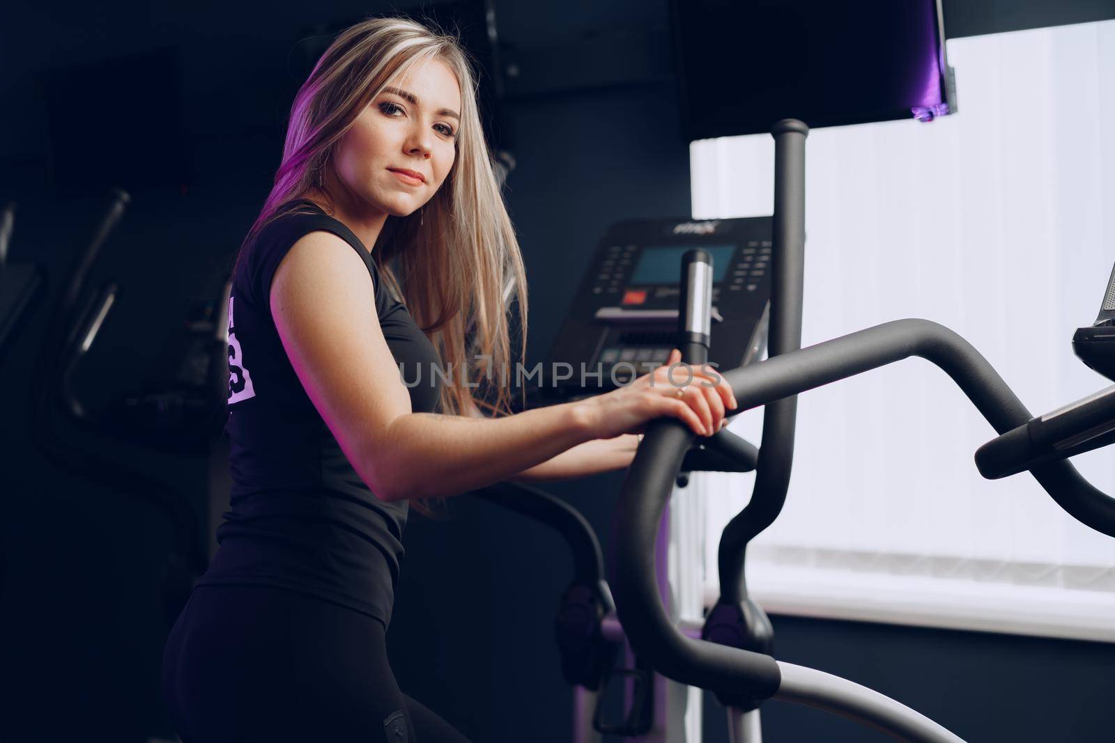 Attractive young blonde woman exercising on cardio training apparatus in dark gym